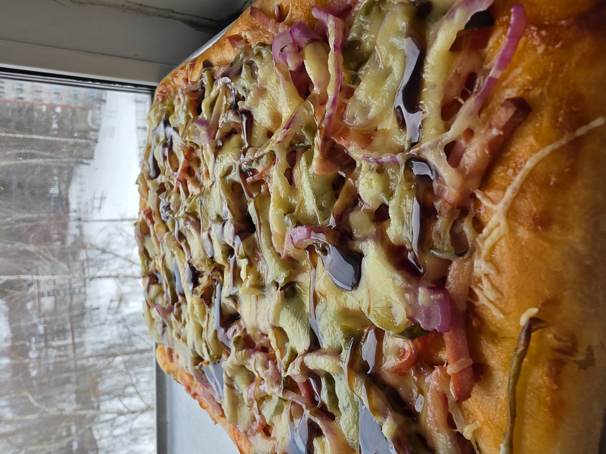 A rectangular pizza with a thick, golden-brown crust occupies the foreground, positioned on a light-colored windowsill. The pizza is topped with melted yellow cheese, sliced red onions, sliced green gherkins or pickles, and visible pieces of red-orange meat, likely ham or bacon. A dark, viscous sauce is unevenly drizzled over the cheese and toppings. The cheese appears recently melted and slightly bubbly.
Beyond the window, an out-of-focus winter cityscape in Chaikovsky, Russia, is visible. The ground is covered in snow. Numerous bare-branched deciduous trees, some with white bark, stand prominently. Multi-story buildings are observable through and behind the trees, indicating an urban or suburban residential area. One distant, light-colored building with multiple windows is centered in the background. The sky is overcast, suggesting a cold, grey day.