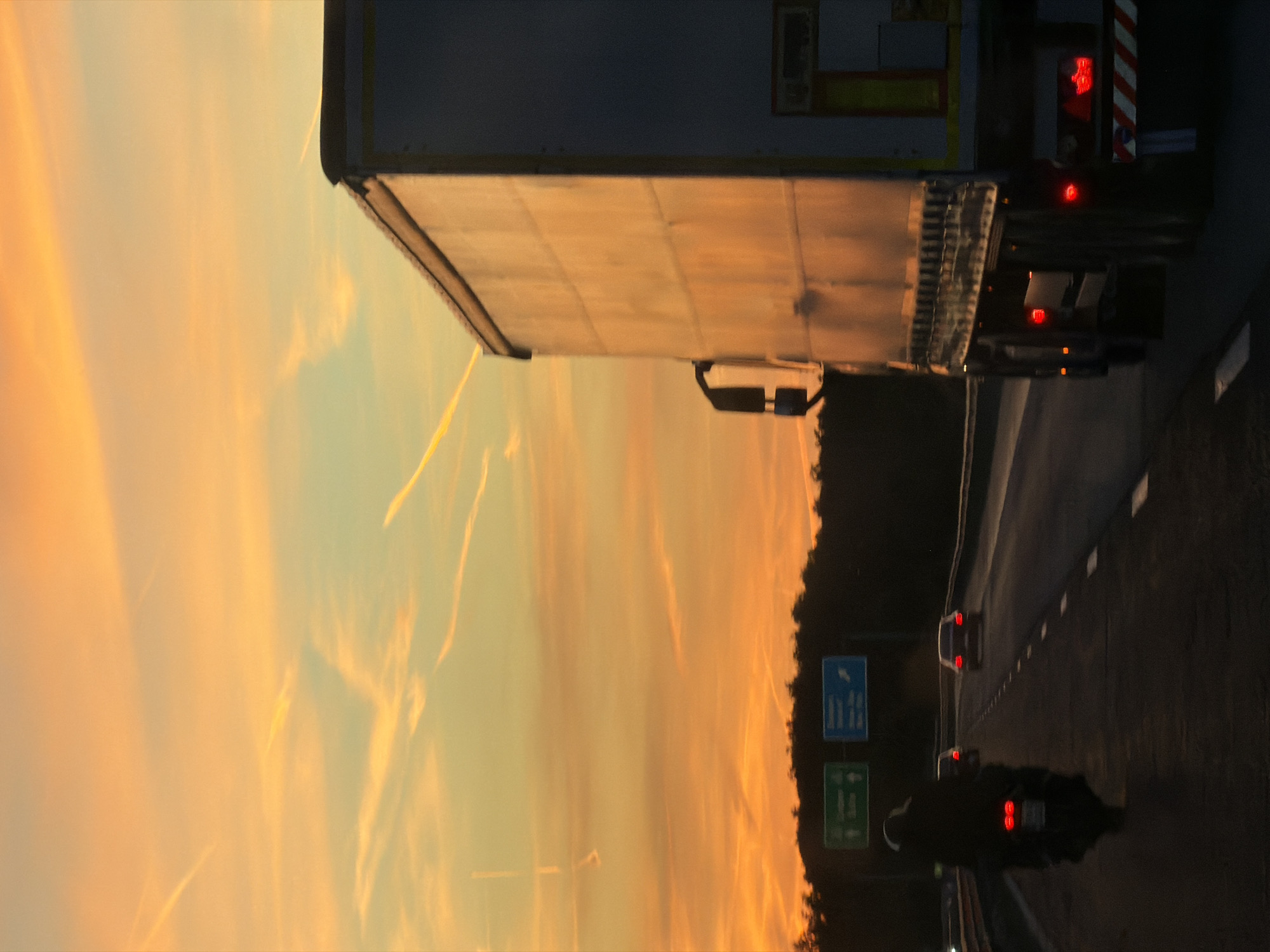 The image depicts a highway scene in Baliovtsi, Bulgaria, at dusk. The sky shows a vibrant orange and yellow sunset with contrails. A large semi-truck occupies the upper portion of the frame, with its trailer illuminated by the setting sun. Below, the highway stretches into the distance, populated by a few cars and a motorcycle, all with their headlights on. Road signs are visible along the roadside. Trees line the edge of the highway.