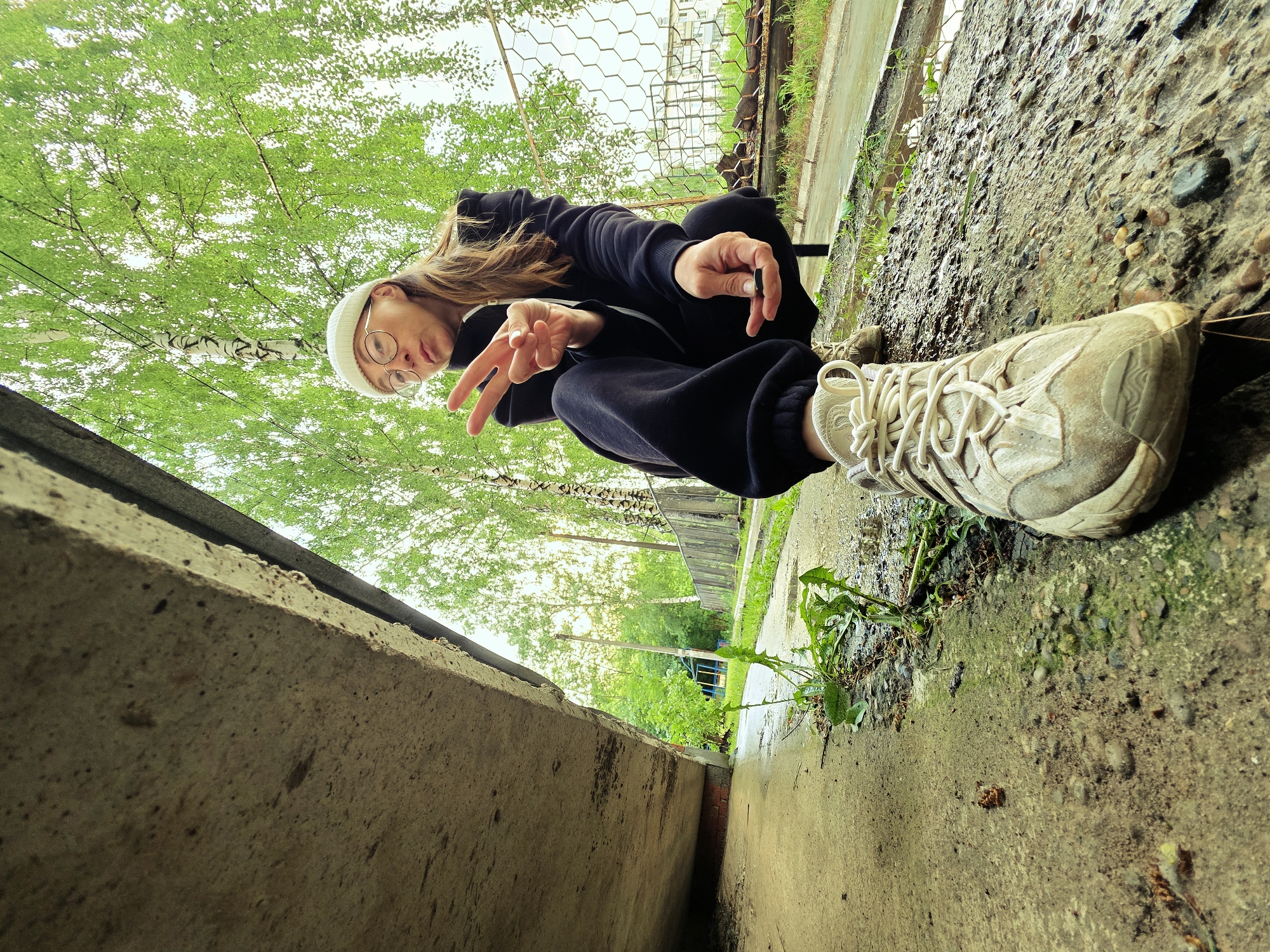 A low-angle shot depicts a person squatting outdoors, looking upwards towards the camera. The individual has long, light brown hair and wears a white beanie, round framed glasses, a dark hooded tracksuit, and light-colored, somewhat worn sneakers. Their left hand forms a peace sign, while the right hand holds a small, dark cylindrical object.
The background is filled with the bright green foliage of trees, particularly noticeable are the white bark of birch trees, creating a vibrant canopy overhead. A chain-link or wire mesh fence is visible behind the person, and in the distance, parts of a pathway or road and a building can be discerned through the greenery.
The foreground consists of rugged concrete structures. On the left, a thick concrete ledge or barrier frames the shot. To the right of the person, the ground is a mix of rough concrete, dirt, gravel, and patches of green weeds, with some areas appearing damp.
The image is captured during daytime, characterized by bright, possibly overcast or diffused light filtering through the trees. The setting suggests an urban or semi-urban outdoor area. The overall impression is casual and relaxed. No visible text is present in the image. The location is Chaikovsky, Russia.