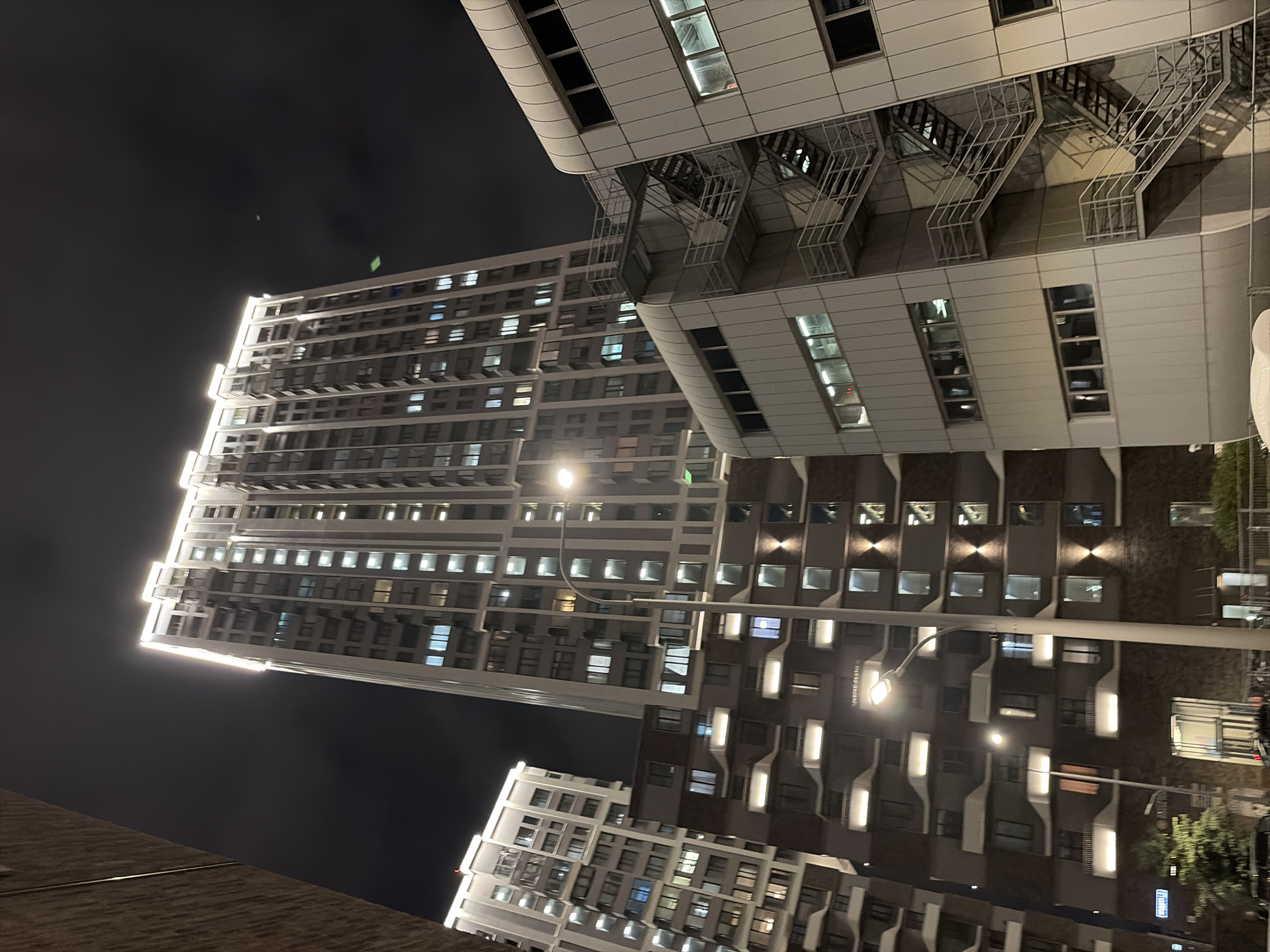 This is a nighttime, eye-level, medium shot of a complex of modern buildings in Moscow, Russia. In the foreground, to the right, is a white, multi-story building with large windows and an external metal staircase. Behind it and to the left is a darker, multi-story building with many illuminated windows and balconies. Further in the background, on the left, is another tall, modern building, and the tallest building in the center is illuminated around its edges. The sky is dark and cloudy.