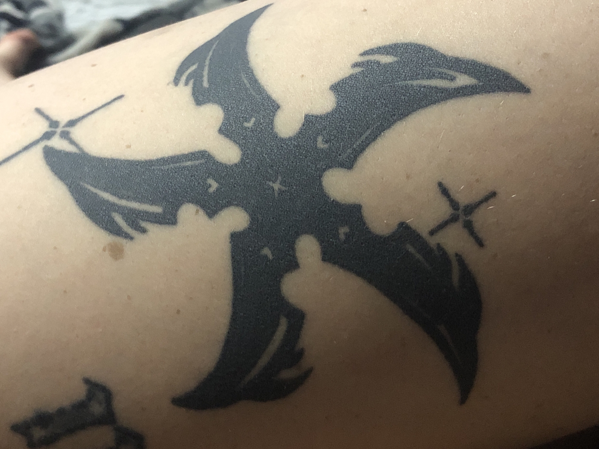 The image shows a tattoo on a person's arm. The tattoo is a black, stylized bat-like figure with sharp edges and wing-like appendages extending from a central body. There are also small, cross-like symbols at the tips of two of the appendages. The tattoo is located on the person's arm. There is another tattoo in the bottom left corner. The arm is fair-skinned with some visible hair and pores. The background is blurry, but appears to be soft grey fabric. No information provided relates to a specific location in Buenos Aires, Argentina.