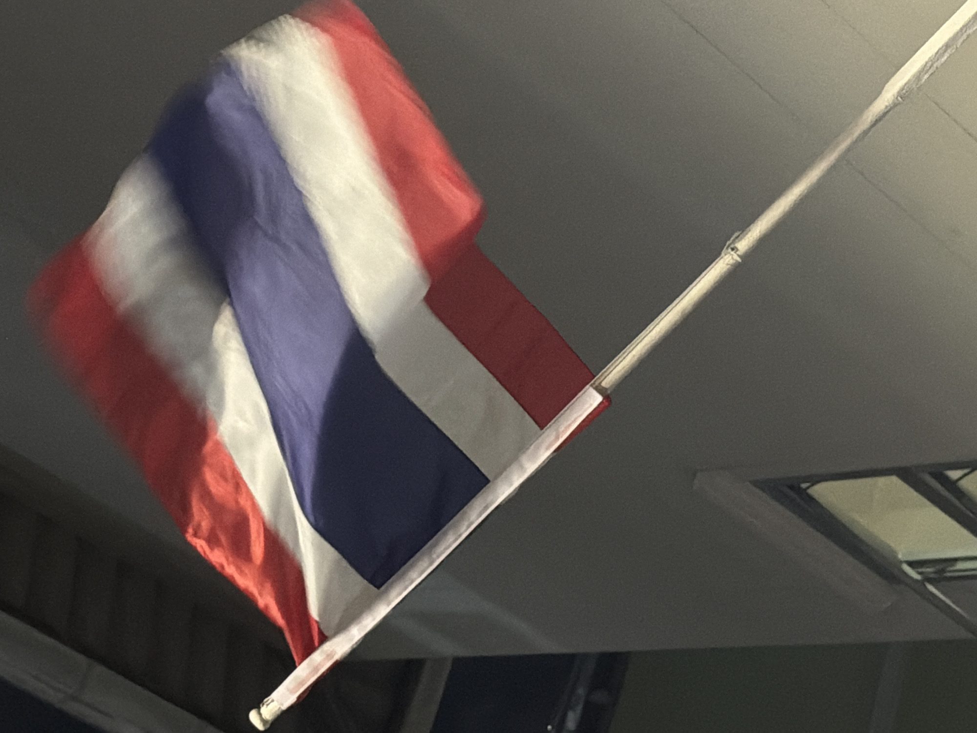 A prominently displayed Thai national flag, characterized by its five horizontal stripes (red, white, wide blue, white, red), is captured in motion, exhibiting significant blur, particularly along its trailing edge, indicating it is actively waving in the wind. It is affixed to a light-colored flagpole, angled upwards from the lower right towards the upper left of the frame. The background consists of a plain, light grey concrete or stucco building facade. A dark-framed window with a faint reflection is partially visible in the lower left section. The scene is illuminated under low-light conditions, likely at night, with artificial light highlighting the flag and a portion of the building. The image is set in an urban environment within Bangkok, Thailand. No people or specific interactions are visible.