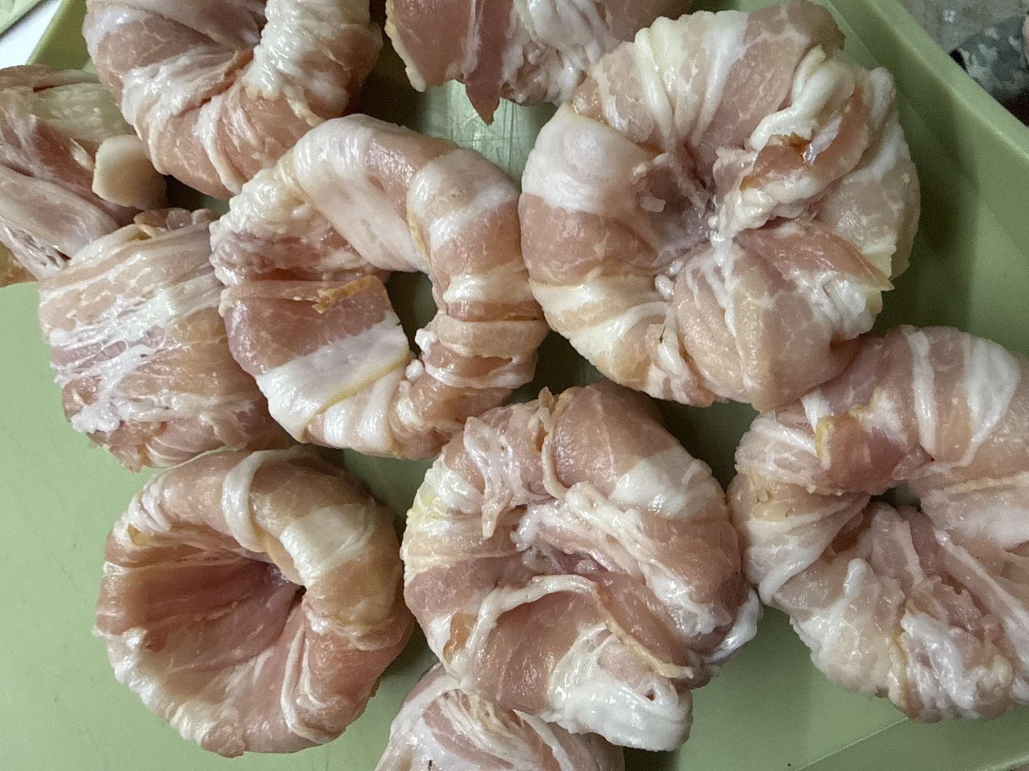 A close-up, high-angle shot shows a light green rectangular tray filled with raw, uncooked bacon-wrapped food items. These items are circular in shape and appear to be small, dense portions of food, possibly scallops or bite-sized pieces of meat, tightly wrapped in strips of raw bacon. The bacon is a mix of pinkish-red lean meat and white fat, arranged in a spiral pattern around each food item, creating a textured surface. Some of the bacon strips have slightly pulled away from the centers of the items, revealing the food within. The tray is placed on a flat surface, and the lighting is bright, casting minimal shadows. The image does not depict any recognizable landmarks or specific details that would allow for the identification of its location in the city of Kamyshin, Russia.
