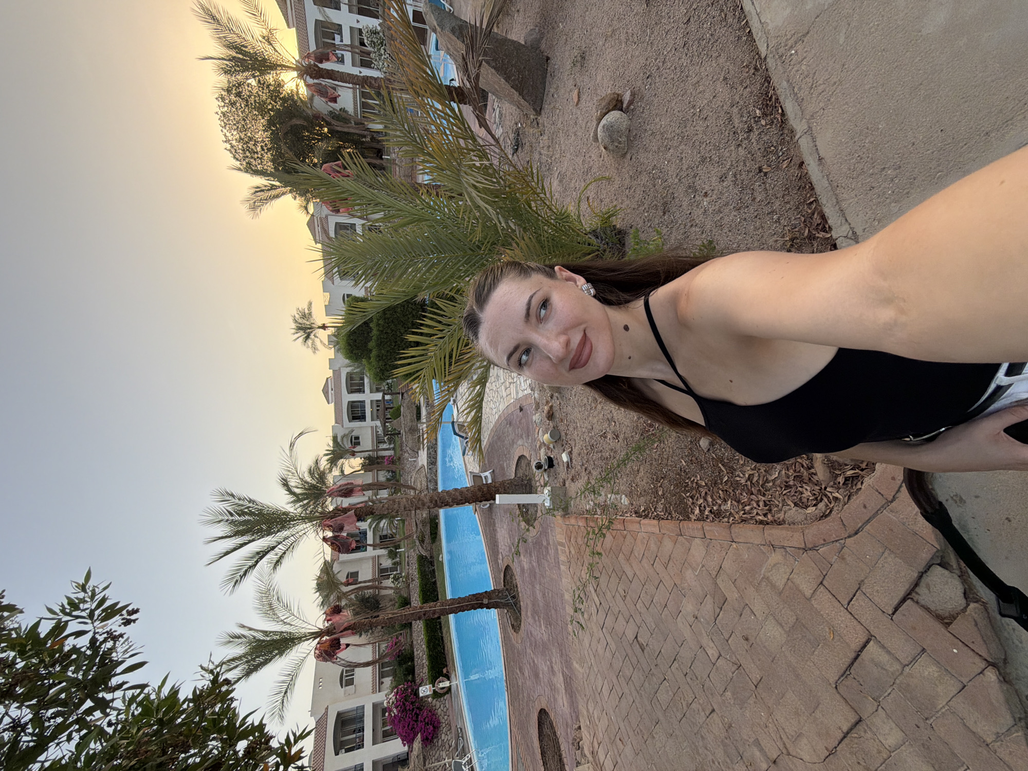 A young woman with long brown hair, wearing a black tank top and white pants, takes a selfie in a resort setting. She has light skin, freckles, and is wearing earrings and makeup. She is standing near a palm tree in a sandy area with a brick pathway to her left. In the background, there is a bright blue swimming pool, more palm trees, and white buildings with red-tiled roofs, indicative of architecture common in Sharm El Sheikh, Egypt. The sky is a pale orange, suggesting it is either sunrise or sunset. A large rock and some smaller stones are visible on the sand. A stroller is partially visible in the lower left corner.