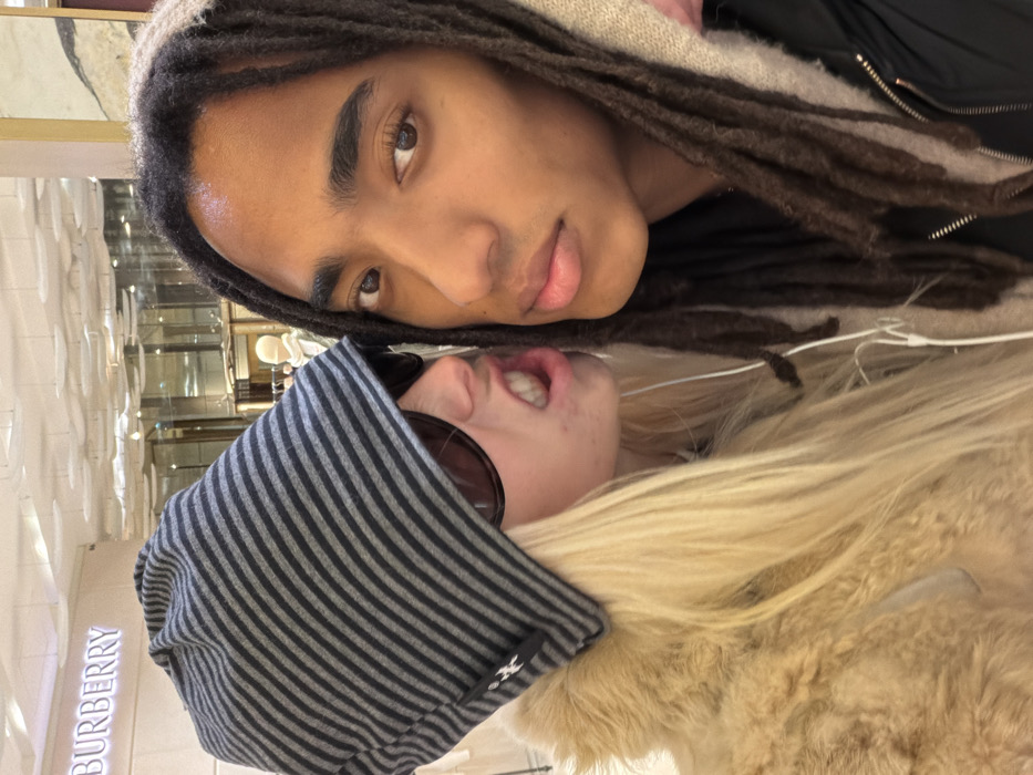 Two people pose closely together. One person has dark skin, dark dreadlocks, and wears a dark top. The other person has light skin, blond hair, and wears a striped hat and sunglasses. They seem to be at a Burberry store, as the store’s signage is visible on the left. The blond-haired person is wearing a furry jacket. Earbuds are hanging around their necks.