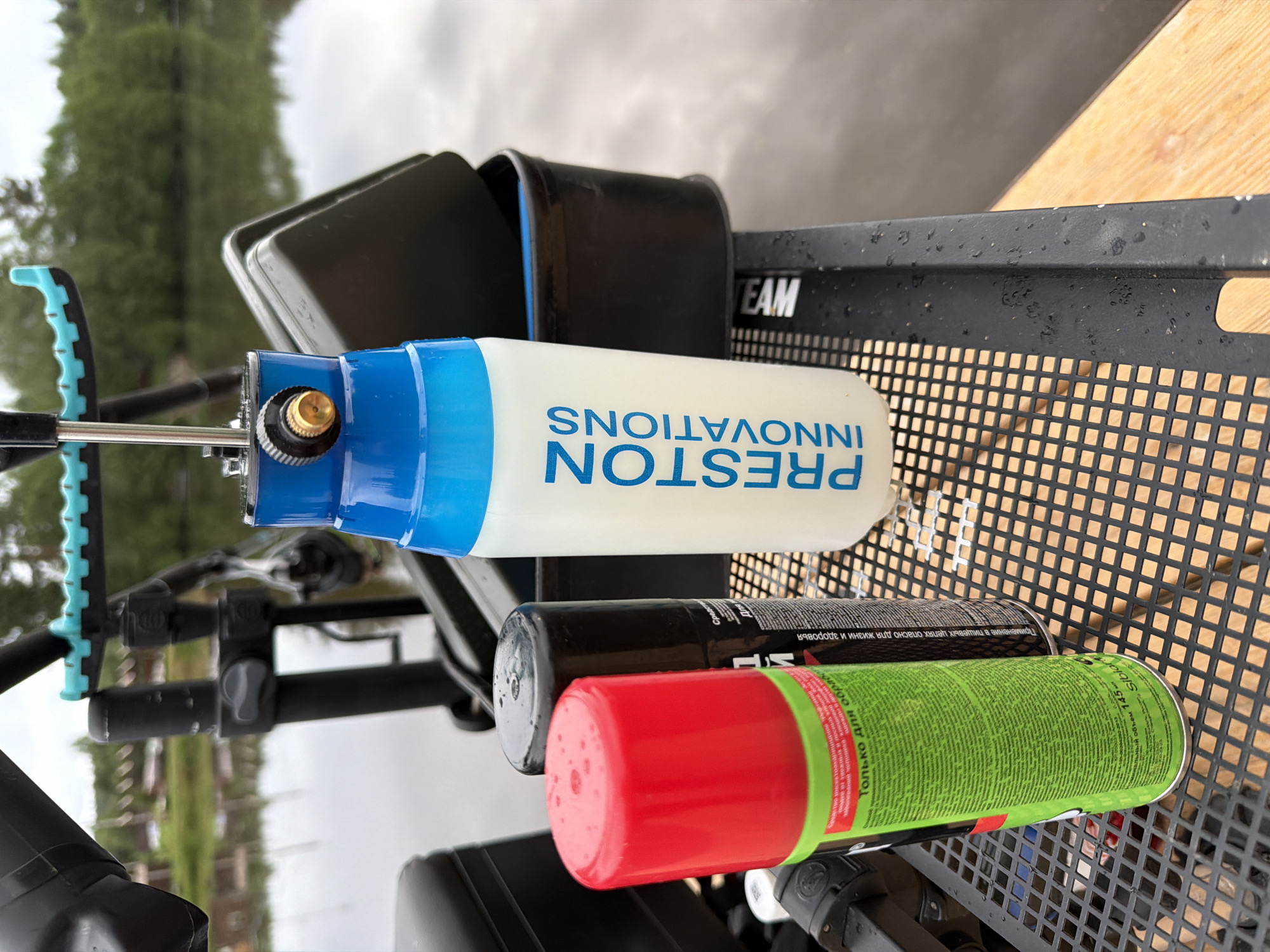 The image shows various items including a tube labeled "PRESTON INNOVATIONS" in a blue and white design, lying on a black metal grate that is part of a "TEAM" branded structure. Below it are two aerosol cans, one with a red top and green body labeled in Russian, and the other black. The background consists of a blurred natural environment with trees and a water body, indicating a location near a lake or river. Based on the OCR text in the image, the aerosol can is from "Siblar" and labeled in Russian indicating it is for "insectoacaricidal" and to be only used for treatment, meaning the photo was possibly taken in the city of Прикамский, Russia.