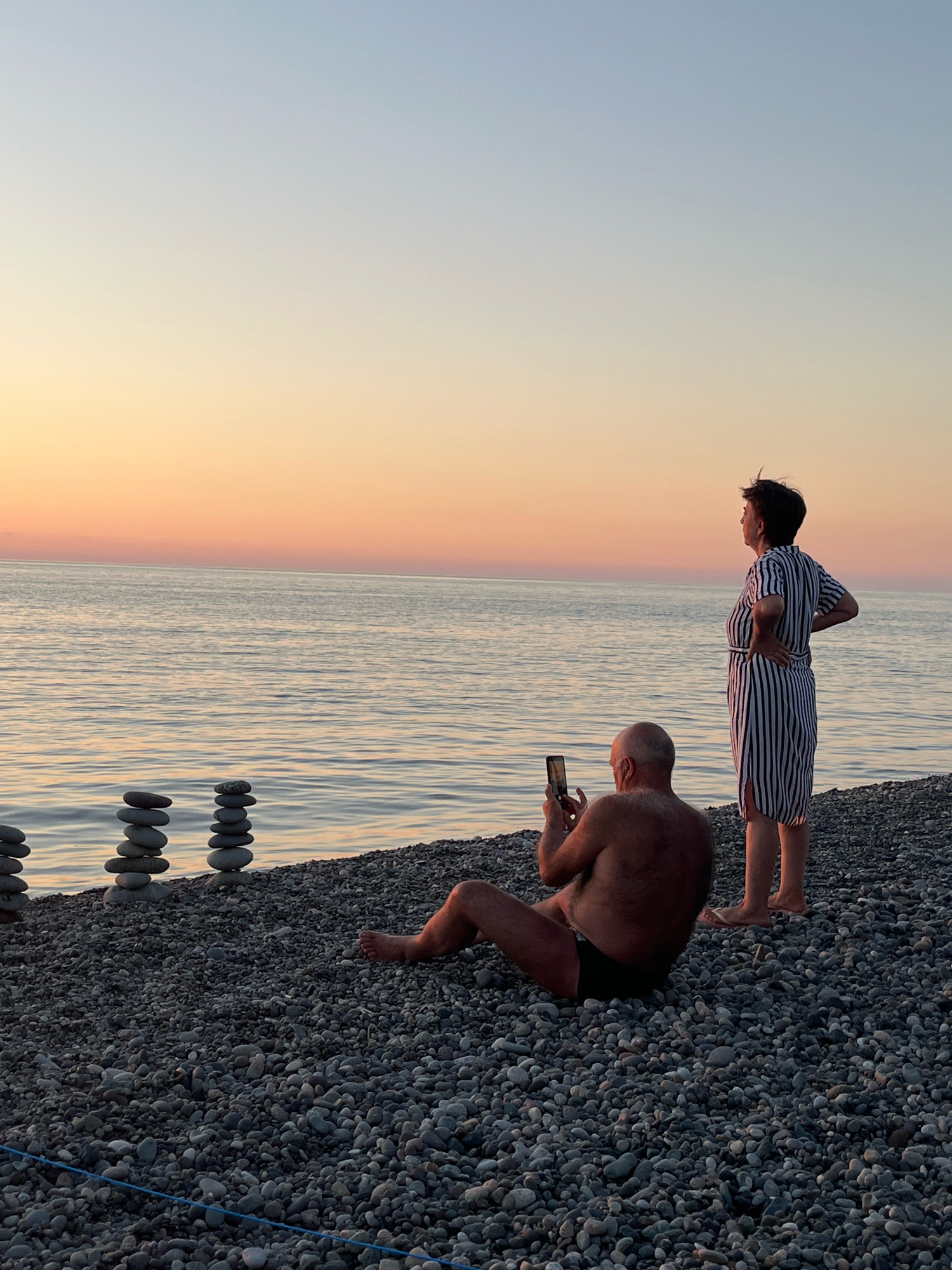 A middle-aged man and woman are standing on a pebble beach at sunset. The man is sitting down, facing away from the camera, and holding a smartphone to take a picture. The woman is standing, facing the sea with her back to the camera, and has one hand on her hip. The sea is calm with gentle waves, and the sky is a gradient of pink and orange hues. In the background, to the left, are three stacks of balanced stones. The scene appears to be on the Black Sea coast near Kobuleti, Georgia.
