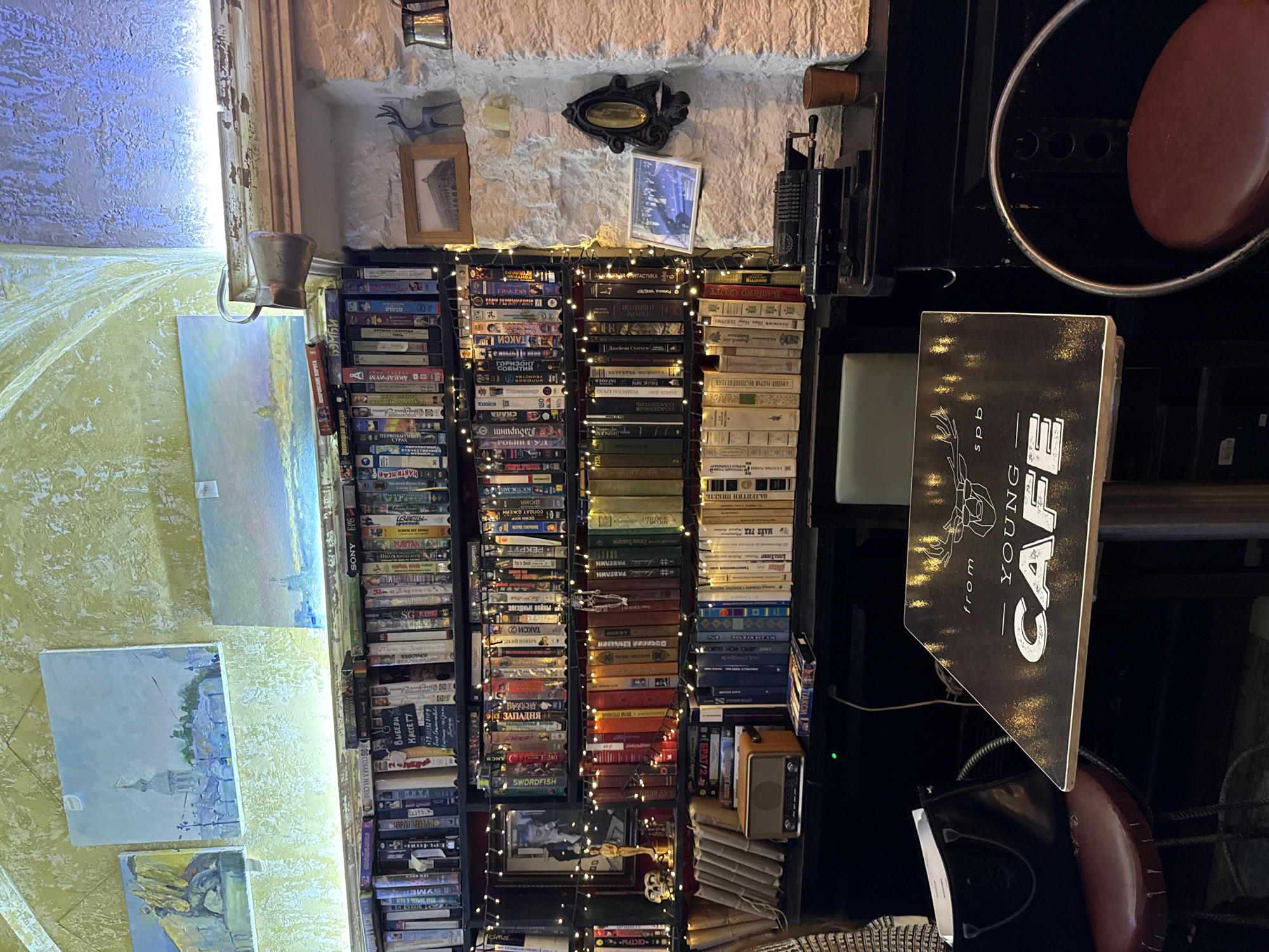 The image depicts the interior of a cafe, likely in Saint Petersburg, Russia, based on the painting style and potential Russian text on some items. The scene is dimly lit, with warm lighting from string lights and overhead lamps illuminating a wall lined with bookshelves. These shelves are filled with a mixture of books and VHS tapes, suggesting a curated collection.
In the foreground, a round table with a dark surface and white lettering that reads "from young cafe spb" is prominently featured. Beside it, a dark handbag rests on a red leather stool. To the right of the table, a vintage-style mechanical device, possibly an adding machine or typewriter, sits on a dark surface, next to a small copper cup. The background reveals a stone wall with decorative elements, including framed pictures and a small metal deer figurine. The ceiling has an arched, textured appearance, painted in a yellowish hue. Several paintings depicting cityscapes are hung on the wall above the bookshelves. The overall atmosphere suggests a cozy and eclectic establishment.