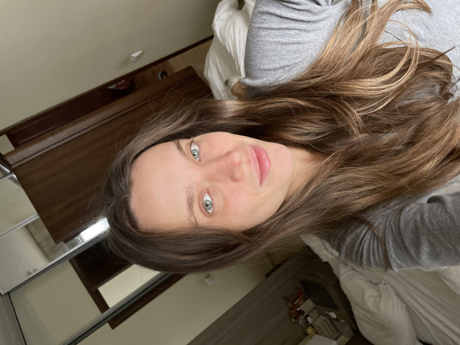 A selfie-style portrait depicts a female subject in an indoor setting in Budva, Montenegro. The subject has long, light brown hair with auburn highlights, green eyes, and a fair complexion with a natural flush. She is wearing a grey, ribbed, long-sleeved shirt and exhibits a slight smile. The background identifies the location as a room, likely a bedroom, with white bedding visible. To the left of the subject, a mirrored wardrobe door reflects a window with blinds and a bedside table containing various objects. To the right, a dark wooden door is partially ajar, revealing an interior space. The scene is evenly lit.
