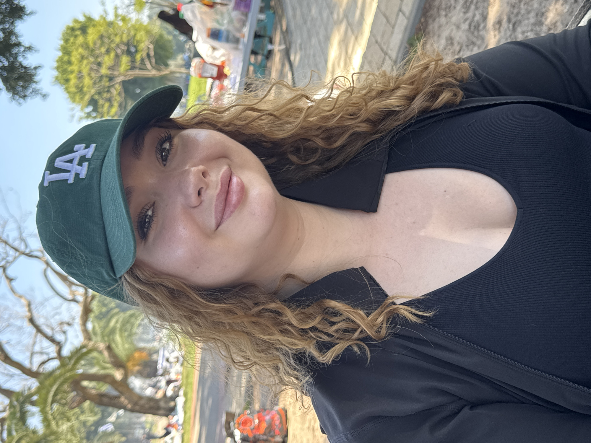 An outdoor portrait captures a young woman smiling gently at the camera in what appears to be a park or recreational area in Ramat Gan, Israel. The woman, with light skin and long, curly blonde/light brown hair, is wearing a dark green baseball cap with a white "LA" logo, a black ribbed top, and a black zip-up jacket. She has subtle makeup, including eyeliner and lip gloss. The image is taken during clear daylight, with bright light illuminating her face. The background is softly blurred, showing green trees against a bright sky, and indistinct forms that suggest picnic tables or stalls with various items, possibly indicating a gathering or event. The overall mood is relaxed and cheerful.