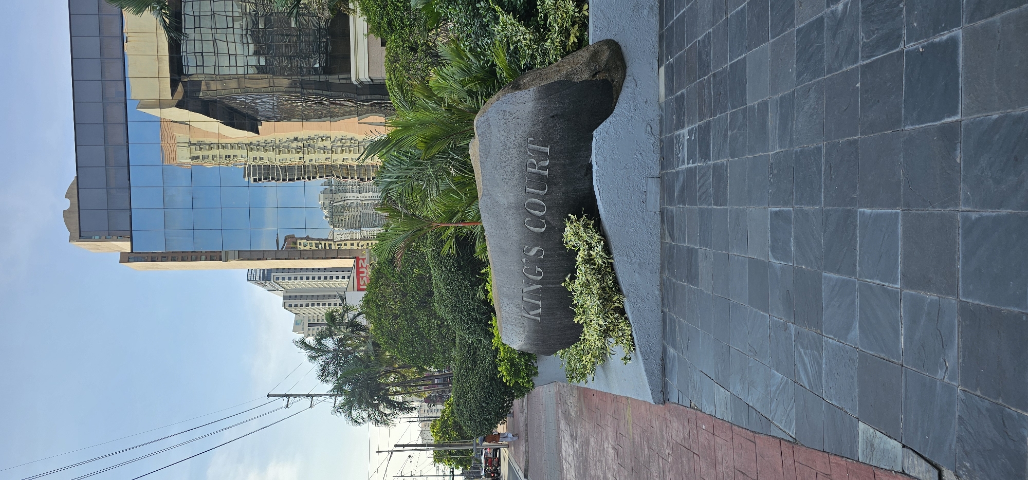A low-angle, full shot captures a stone marker with "KING'S COURT" carved into it, set against a backdrop of lush greenery and modern high-rise buildings in Makati City, Philippines. The marker rests on a grey concrete base, surrounded by low-lying green and variegated plants. To the left of the marker, a sidewalk paved with red bricks leads towards the street. In the distance, several glass-facade skyscrapers, characteristic of Makati's central business district, reflect the blue sky and surrounding structures. A single person, dressed in white, walks on the sidewalk. Utility poles and power lines are visible on the left side of the frame, indicating an urban environment. The foreground features a tiled walkway made of dark grey square stones.