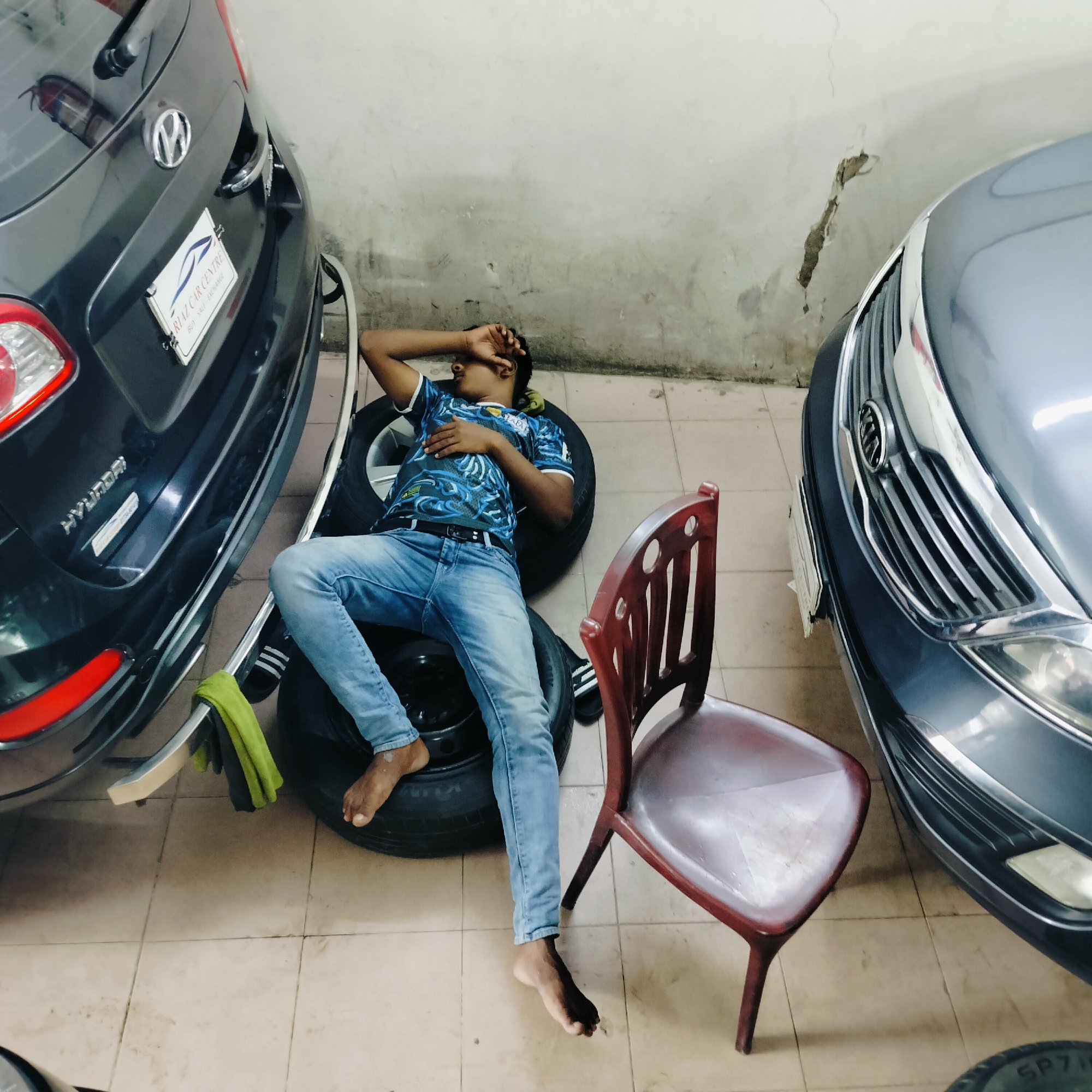In an indoor setting, likely a car workshop or garage in Dhaka, Bangladesh, a young male appears to be asleep. He is lying on a stack of tires, with his head resting on one and his body extended across another. The individual is wearing a blue and black patterned t-shirt, blue jeans, and black and white striped sandals. One hand is covering his face, obscuring it from view.
Adjacent to the tires is a maroon plastic chair, positioned near the feet of the sleeping individual. To the left of the frame, the rear of a dark gray car is visible, featuring a license plate that reads "RIZ CAR CENTRE BUY SELL EXCHANGE." A green cloth is draped over a silver handle or bar on the car. To the right, the front end of a light gray car is partially visible, including its grille and headlights. The floor is tiled in a neutral beige color, with some visible dirt and debris. The background consists of a textured concrete wall, showing signs of wear and cracking.