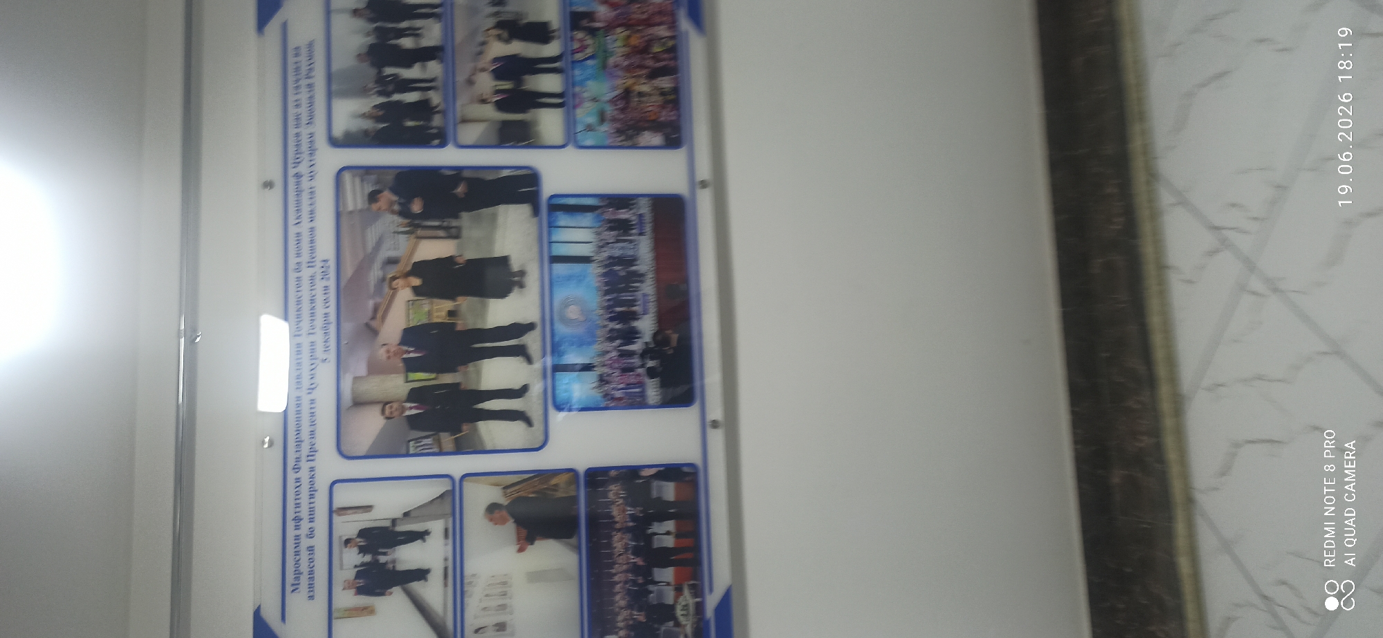 The image displays a wall with a framed collage of photographs. The collage is topped with text in a foreign language, likely Cyrillic or a similar script, which reads "Marosimlar's opening ceremony of the State Philharmonic of Tajikistan named after Abuabdullo Rudaki, President of the Republic of Tajikistan, Emomali Rahmon, on December 5, 2024." The photographs depict formal events with people in suits and traditional attire. One image shows two men walking down a hallway. Another shows three people, two men in suits and a woman in a dark dress, standing in a room. A third photo captures a group of people in suits attending what appears to be a formal ceremony, with a banner or screen in the background. Another image shows a gathering of many people on a stage or platform, some in traditional clothing. The bottom right image is blurry and appears to depict a colorful performance or cultural event. The overall impression is a display commemorating a significant event or series of events, possibly related to a cultural or governmental occasion. The location is not specified beyond a potential reference to Tajikistan in the text.