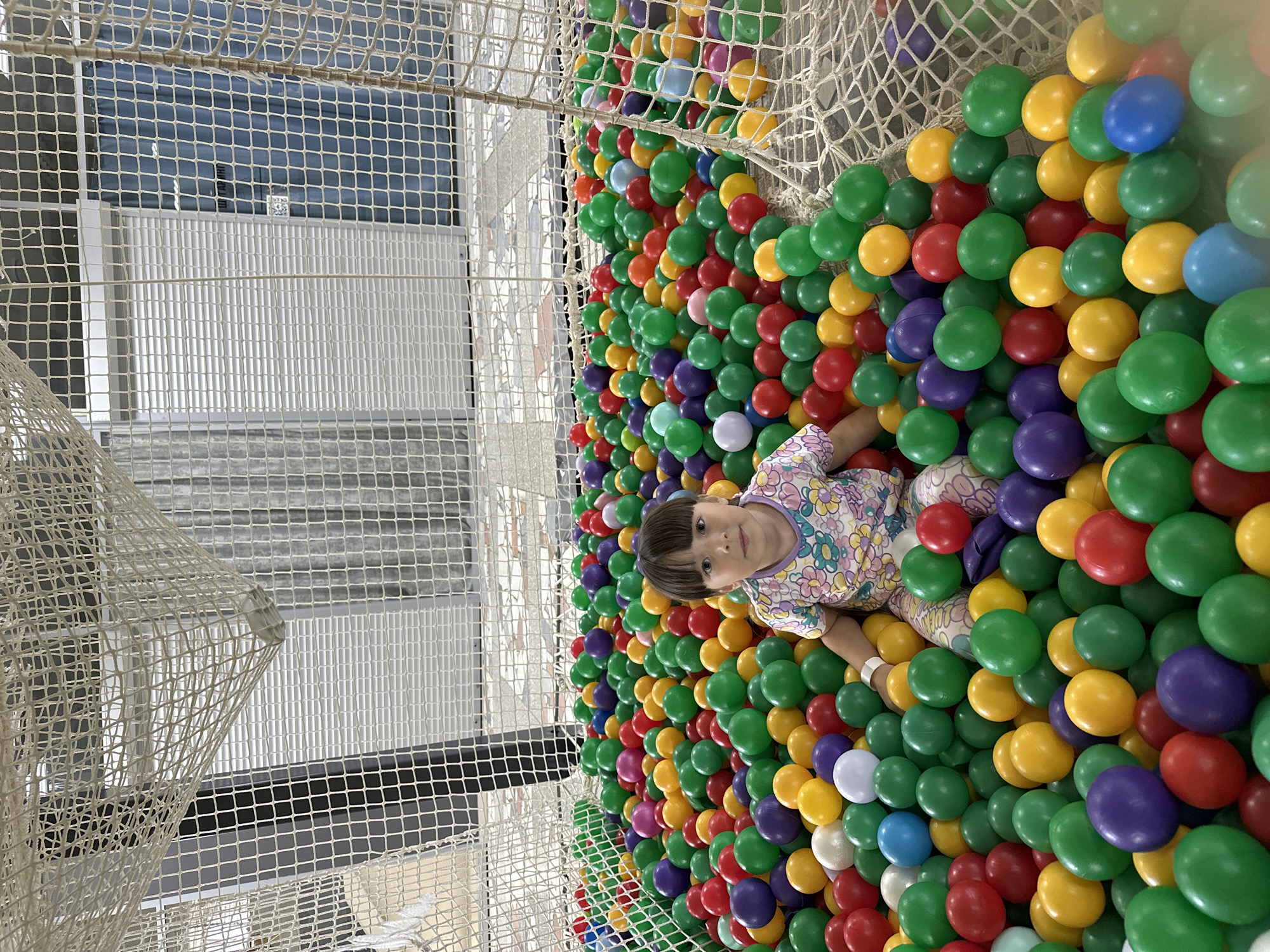 A young child, approximately 2-4 years old, with dark hair and bangs, sits centrally within a large, multi-colored ball pit in an indoor recreational facility in Chaikovsky, Russia. The child wears a light-colored, short-sleeved t-shirt adorned with a floral pattern in shades of pink, yellow, blue, and green, and a white wristband on the right wrist. The ball pit is densely filled with thousands of plastic balls in red, green, yellow, purple, blue, and white, reaching to the child's chest level while seated. The entire ball pit is enclosed by a light-colored, coarse mesh netting. Through the netting in the background, interior building elements are visible, including white walls, white slatted roll-down shutters, and a segment of a dark blue curtain. A light-colored tiled floor is also visible outside the netted enclosure. The child looks directly at the camera with a calm, neutral expression.