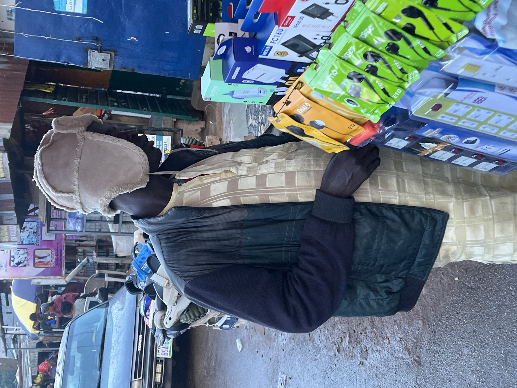 An outdoor market scene in Gura Top, Nigeria, features a man in a light-colored, textured hat and a dark jacket over a patterned tunic. He stands near a display of electronics accessories, including earphones in yellow and green packaging, power adapters in blue and green boxes, and multi-plug adapters. In the background, a gray car with a license plate that reads "BSA 6" is parked. Further back, a stall with yellow and blue umbrellas is visible, along with various other market structures and people browsing. The ground appears to be wet and uneven, suggesting it may have recently rained.