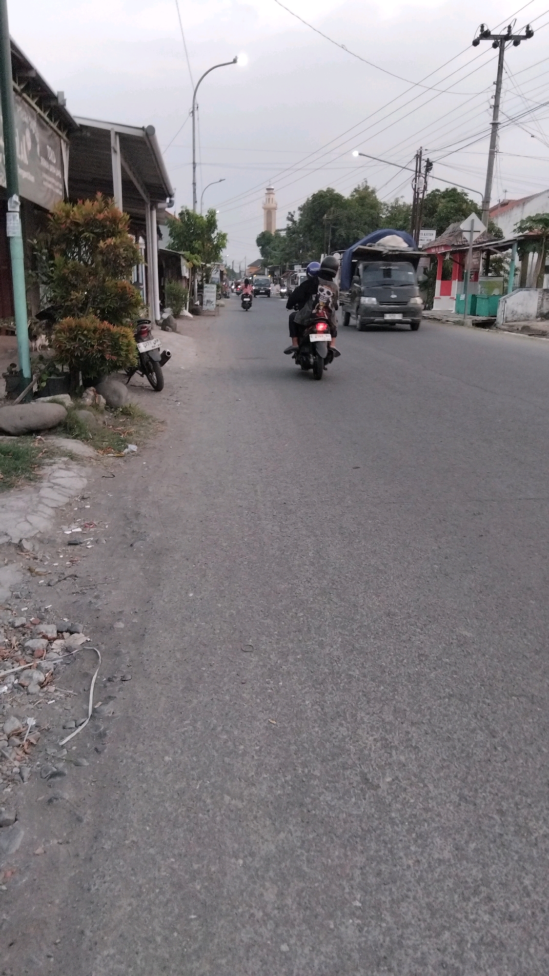 The image depicts a street scene in Tegal, Indonesia, during the daytime under an overcast sky. The road is paved and appears to be a two-lane thoroughfare. On the left side of the road, there are several low-rise buildings with awnings, suggesting a commercial or residential area. Some vegetation, including small trees and bushes, is visible along the roadside. Several parked motorcycles are present.
In the middle of the road, a dark-colored truck with a blue tarp covering its load is moving away from the viewer. Ahead of the truck, two motorcycles are traveling in the same direction. The lead motorcycle carries two individuals, both wearing helmets. The motorcycle behind it also has two occupants, also wearing helmets. Further down the road, other vehicles, including cars and motorcycles, are visible in the distance. A tall, light-colored minaret, likely part of a mosque, is prominent in the background. Utility poles with electrical wires line the right side of the street. The ground on the left side of the road is unpaved and shows debris, including rocks and what appears to be a piece of white plastic.