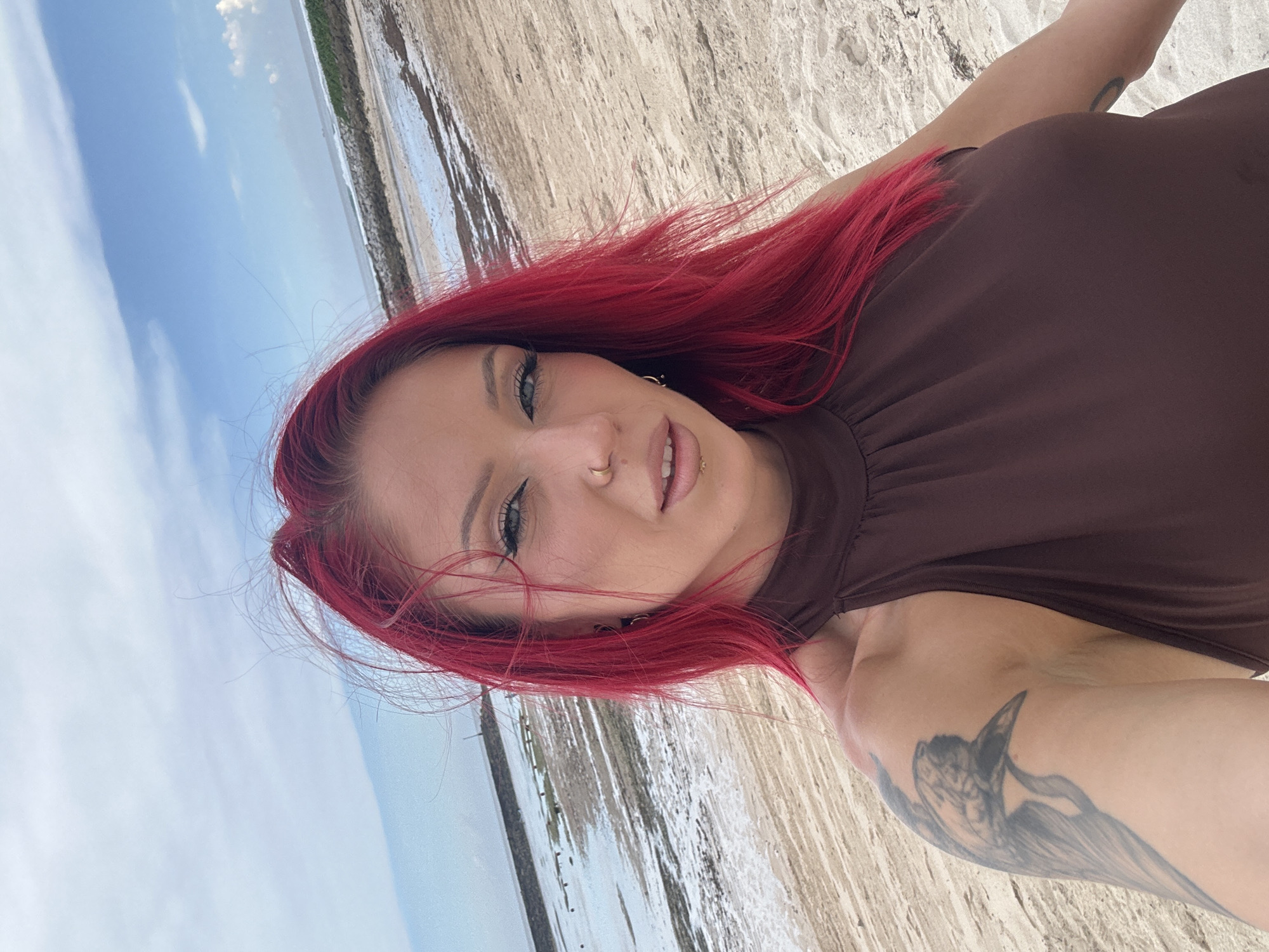 A selfie photograph shows a woman with red hair wearing a brown top. She has a nose ring and a tattoo on her arm. The background appears to be a beach with the ocean and sky visible. Based on the prompt, this is in Nusa Dua, Indonesia.