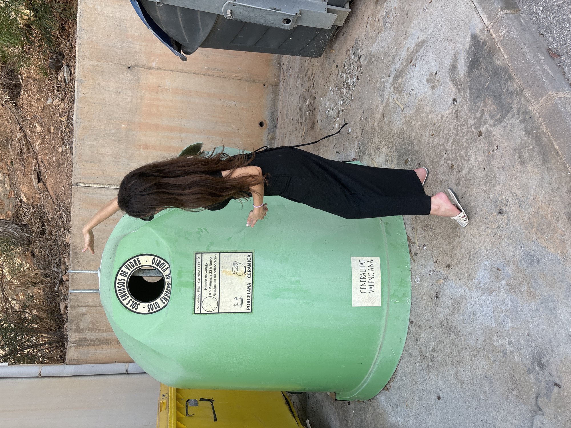 A woman with long, dark hair stands with her back to the camera, facing a large, green glass recycling bin in an outdoor urban setting. She wears a long, black dress and white sandals. Her right arm is raised, with her hand near the top of the bin, and her left hand is resting on the side of the bin. The recycling bin has text in a circular logo at the top and a rectangular label below that indicates it is for porcelain and ceramic. Another label at the bottom of the bin reads "GENERALITAT VALENCIANA". Adjacent to the green bin, to its left, is a yellow recycling bin, and to its right, a large grey dumpster. The ground is paved with concrete, and there is a concrete wall and vegetation in the background. The scene appears to be in daylight.