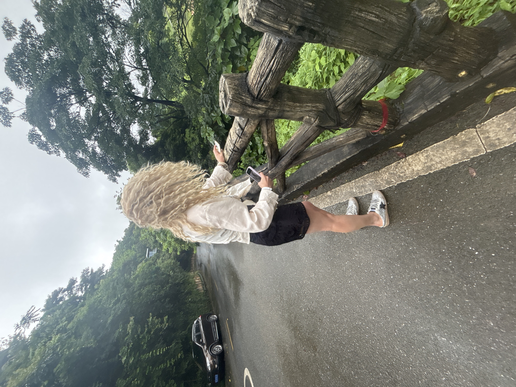 A woman with long, curly blonde hair stands on a wet asphalt road, looking away from the camera. She is wearing a light-colored blouse and a black skirt, with sparkly silver sneakers. She is holding a smartphone in her right hand and reaching out with her left hand towards a wooden fence that runs alongside the road. The fence is made of rough, weathered logs. The road is lined with lush green foliage and trees, suggesting an outdoor, possibly mountainous or park-like setting. A dark brown SUV is parked on the left side of the road, further down. The sky is overcast, indicating a cloudy day. The scene appears to be in a humid environment, given the wetness of the road.