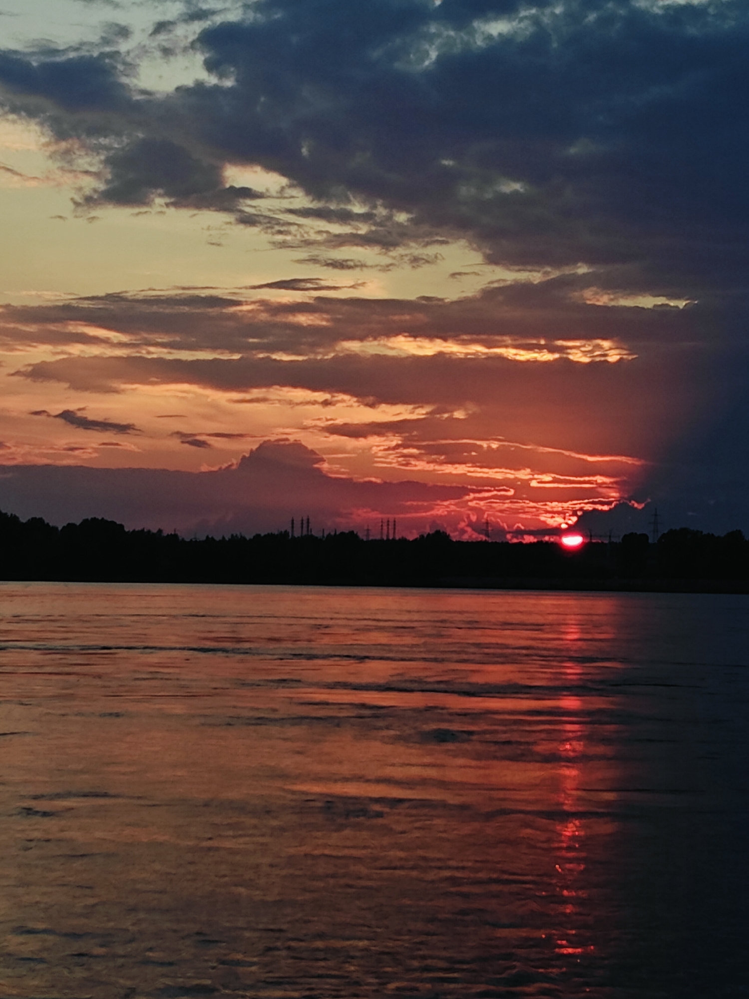 The image depicts a sunset over a body of water, likely the Kama River, as seen from Chaikovsky, Russia. The sky is filled with dramatic clouds, ranging from dark gray to vibrant orange and red, illuminated by the setting sun. The sun itself is partially obscured by clouds, casting a fiery glow across the scene.
In the foreground, the water reflects the colors of the sky, with streaks of orange and red shimmering on its surface. The horizon is lined with a dark silhouette of trees and what appear to be industrial structures, possibly power lines and chimneys, which are characteristic of the city's landscape.
The overall impression is one of natural beauty juxtaposed with human industrial presence, common in many Russian cities situated along major rivers. The scene is tranquil, captured during the twilight hours.