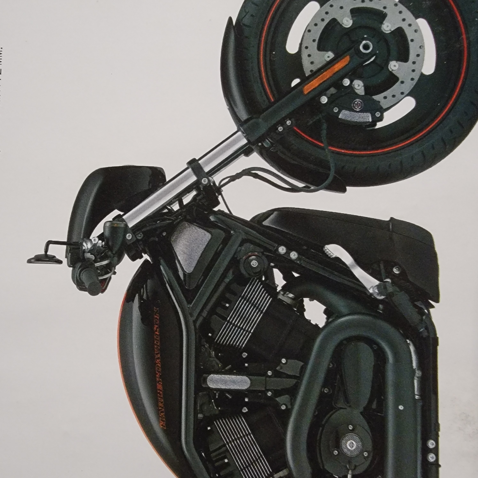 The image shows a close-up, side view of the front section of a black Harley-Davidson motorcycle. The motorcycle is stationary against a plain white background, with no city or environmental context.
Key features visible include:
* **Front Wheel and Brake:** The front wheel is black with a red accent stripe along the rim. The brake disc is silver with multiple perforations and a black brake caliper is attached. An orange reflector is visible on the front fork.
* **Engine and Frame:** The prominent V-twin engine is black, with silver fins on the cylinders. The motorcycle's frame and exhaust pipes are also black. A textured, silver insert is visible on the side of the engine cover.
* **Handlebars and Mirrors:** The handlebars are black, and a single black rearview mirror is mounted on the left side.
* **Fuel Tank:** A portion of the fuel tank is visible at the top left, also black with an orange pinstripe and the "HARLEY-DAVIDSON" logo in orange lettering.
* **Other Details:** Various metallic components, bolts, and wiring are visible throughout the motorcycle's front assembly.
There are no people, animals, or any indication of a specific location or city. The lighting is even, suggesting a studio or controlled environment for product photography.