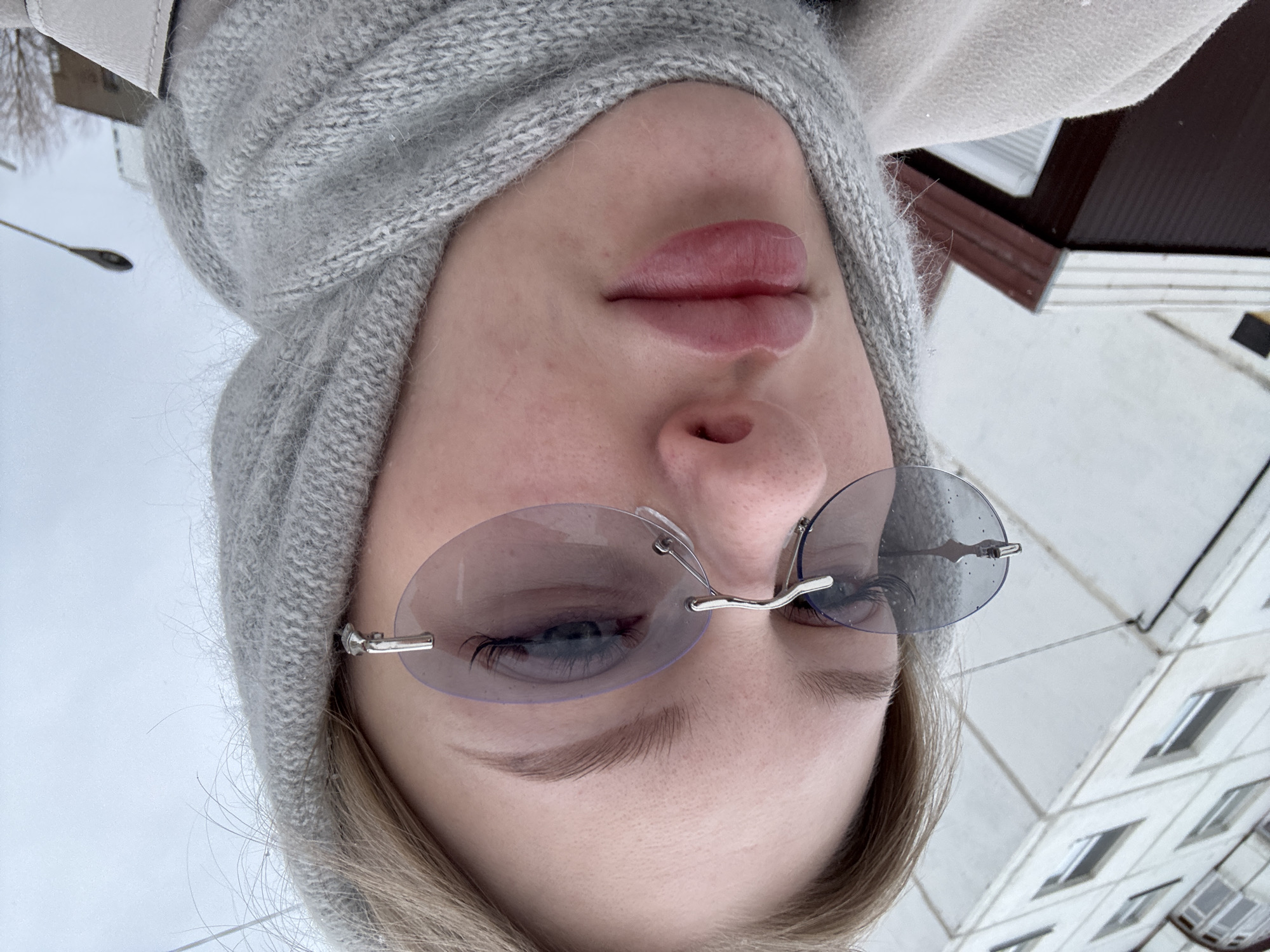 An upward-angled, inverted close-up shot focuses on a person's face against an outdoor urban backdrop, identified as Perm, Russia. The subject, likely female, has light-colored hair partially covered by a grey knitted beanie. Her full, neutral-toned lips are prominent in the center, and a portion of her nose is visible. She wears rimless, oval-shaped sunglasses with light, possibly blue or purple-tinted lenses, through which her blue eyes and long eyelashes are faintly discernible. Her eyebrows are well-defined.
The background reveals a grey, overcast sky, suggesting daylight but potentially cold or dreary weather. To the left, a street light pole stands against the sky. To the right, part of a multi-story building is visible, featuring a light-colored, possibly concrete or stucco facade with a grid pattern and several windows, along with a dark roofline. The warm attire (beanie) supports the notion of a cool climate. The person appears to be looking downwards towards the camera, possibly taking a selfie. No visible text is present in the image.