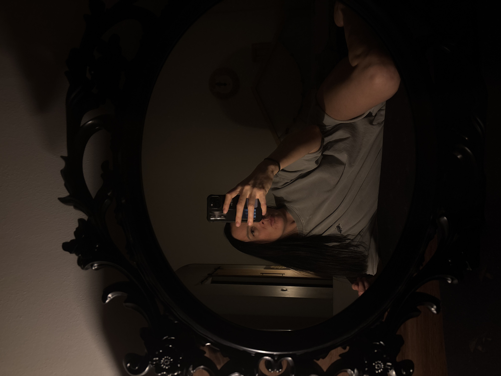 A person is reflected in an ornate, black oval mirror. The reflection shows the person holding a smartphone, presumably taking the picture. The person has long, dark hair and is wearing a light gray sweatshirt with a small logo visible. The background behind the person appears to be a dimly lit room. The specific location in the city of Chaikovsky, Russia, cannot be determined from the image alone.