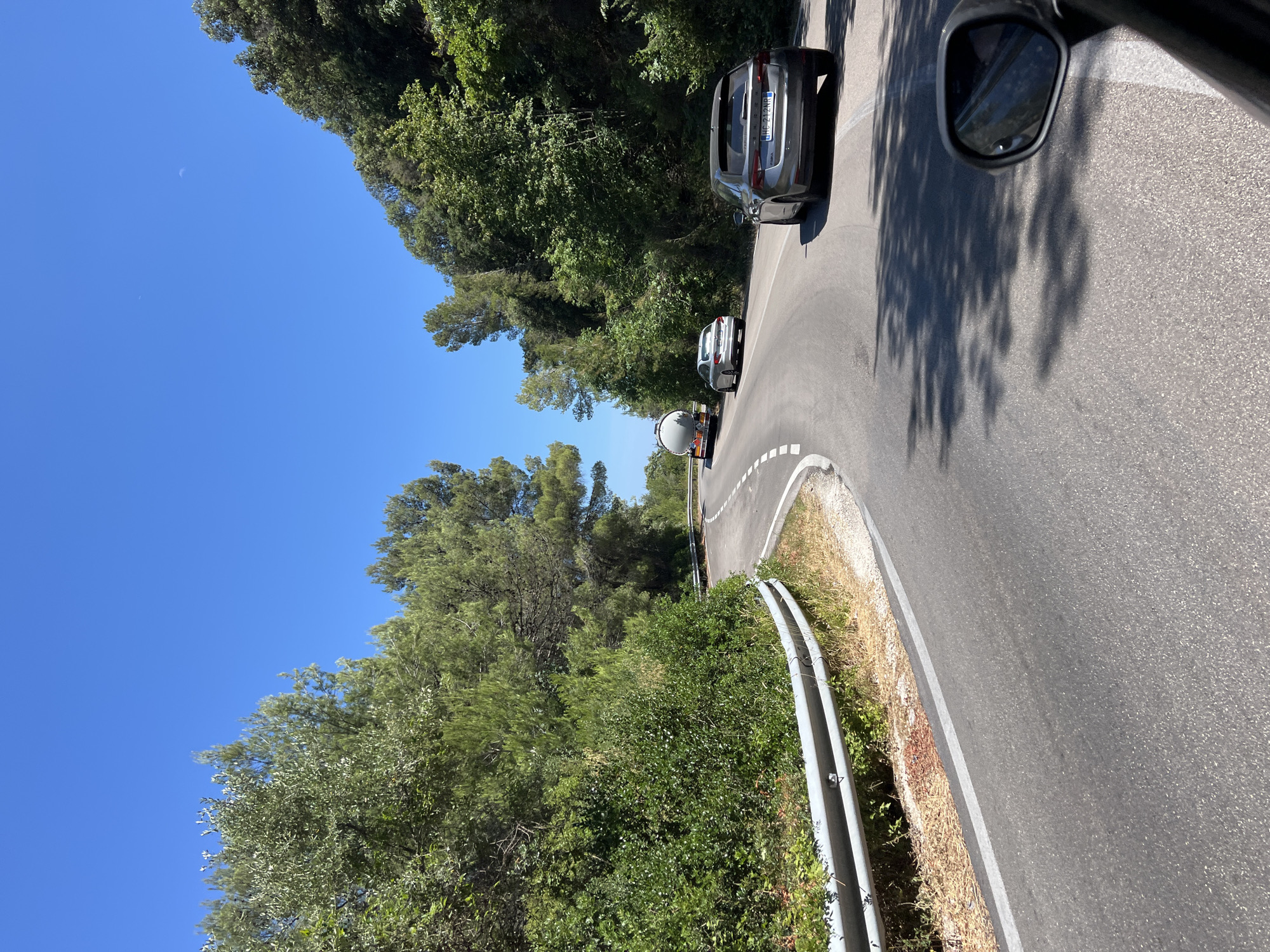 The image shows a winding road in a wooded area, likely in the vicinity of Mattinata, Italy. A silver sedan is in the foreground, driving on the road. Further ahead, another silver sedan is visible, as well as a white tanker truck. The road is bordered by lush green trees and bushes, with a metal guardrail on the left side of the road. The sky is a clear blue with a few faint white wisps. The overall scene suggests a scenic drive through a natural landscape.