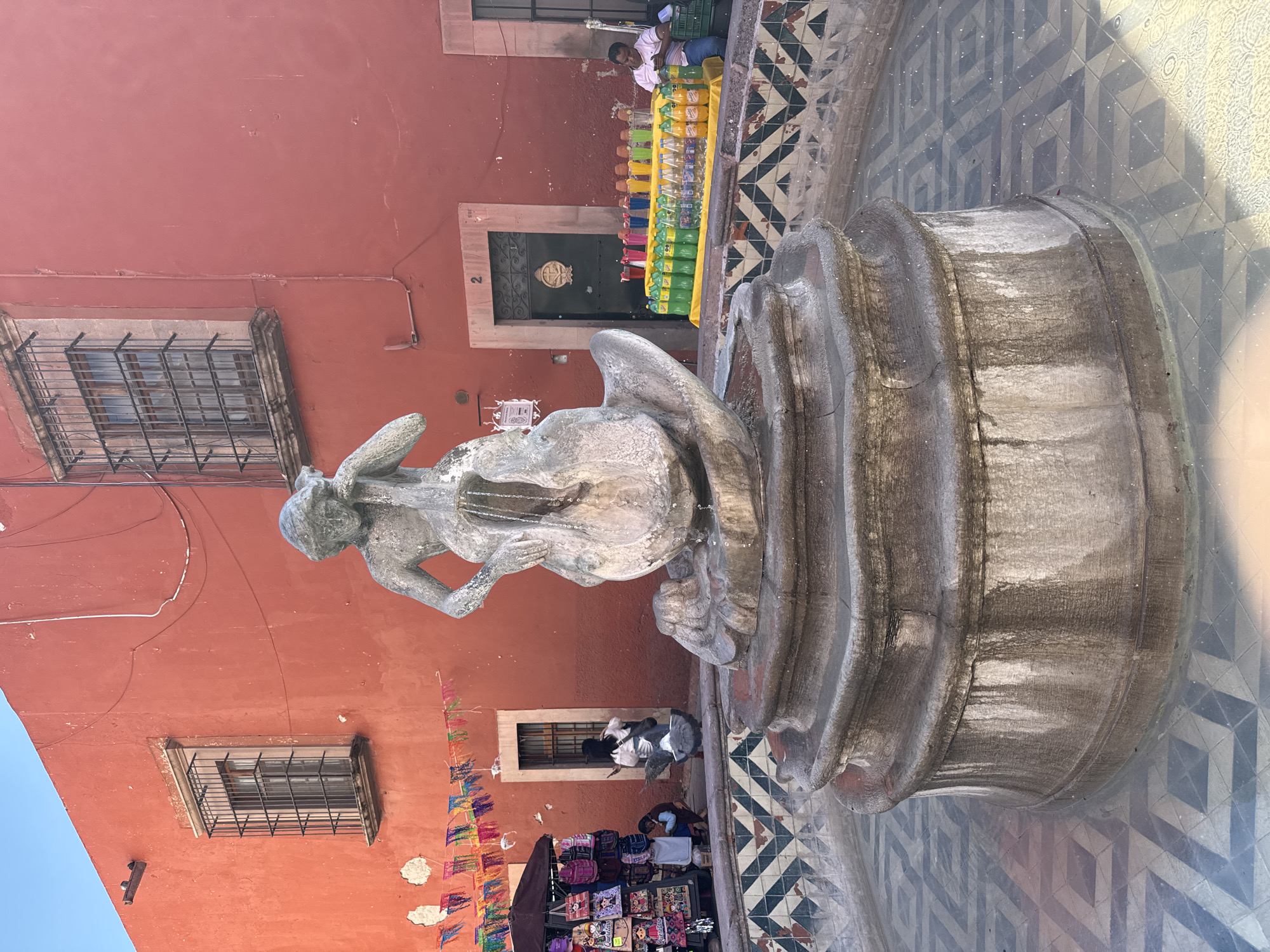 The image shows a sculpture atop a water fountain in a public square. The statue appears to depict a woman playing a stringed instrument. The fountain rests on a tiled surface with a geometric pattern. Behind the fountain is a red-painted building with barred windows and a door with a number "2" on it. A person is selling bottled drinks from a table along the building. Banners are strung along the building in another area, where another person is seen selling souvenir-type products from a pop-up stall. The location is the city of Querétaro, Mexico.