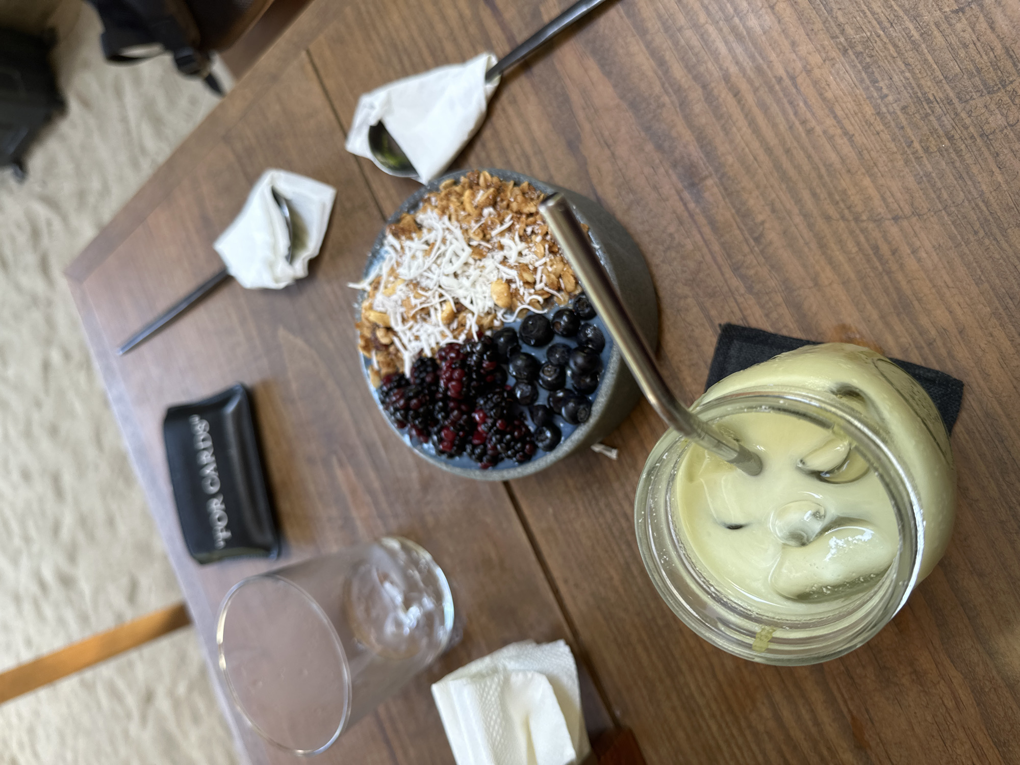 The image depicts a tabletop scene, likely in a café, showcasing an assortment of items suggestive of a meal or refreshments. A bowl filled with a mixture of granola, shredded coconut, blueberries, and blackberries is central to the composition. A metal spoon rests within the bowl. Adjacent to the bowl is a glass jar containing a greenish beverage, possibly matcha or a similar blended drink, with a straw inserted. A clear glass, likely for water or another beverage, is nearby, accompanied by a stack of folded white napkins. There's also a small black cardholder labeled "FOR CARDS." The scene is set against a wooden tabletop with visible grain. The lighting suggests a natural or well-lit indoor environment. The location is not discernible based on the image alone; it is not possible to state the location as Puerto Escondido, Mexico, with certainty.