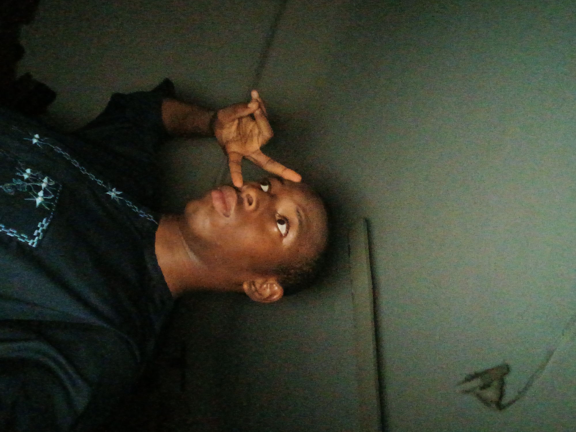 A person is lying on what appears to be a wall in what appears to be a dark-lit room. The person is making a gesture with their right hand that appears to show 3 fingers pointing upward. The person is wearing a dark shirt with what looks like a chain pattern on the chest area. Location: Lagos, Nigeria.