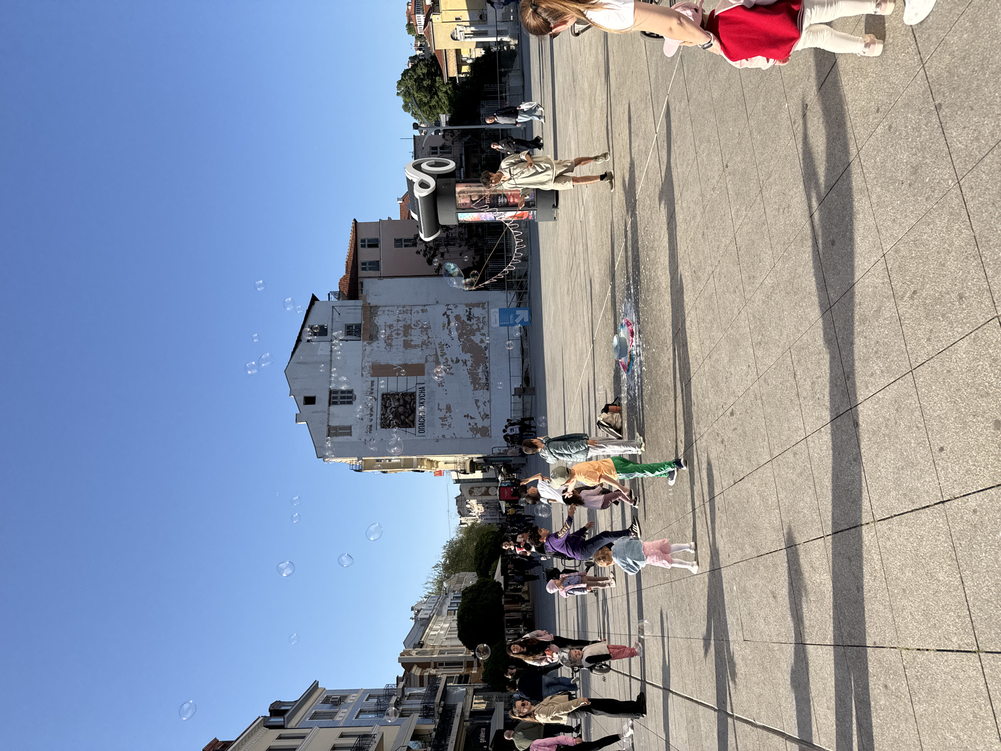 This media file captures a lively scene in a public square in Plovdiv, Bulgaria, during what appears to be late afternoon, given the long shadows cast across the paved ground by the low-angled sun. The sky is a clear, bright blue, indicating sunny weather.
The primary activity depicted is a street performer operating a bubble machine, filling the air with numerous iridescent bubbles. A man in light clothing and shorts stands near a black street advertising pillar, holding the large wands used for creating the bubbles. Numerous people, including adults and many children, are gathered in the square, seemingly enjoying the spectacle. Children are particularly engaged, watching and reaching for the floating bubbles. In the foreground on the right, an adult holds a young child dressed in a red top and white pants. Other groups of people are scattered throughout the square.
The setting consists of a spacious public square paved with light-colored tiles. Various buildings, ranging from older structures with faded paint to more modern ones, line the edges of the square and recede into the background. On a large, distressed white building on the left, visible Cyrillic text includes phrases like "ОПАСНО ВКУСНА" (OPASNO VKUSNA, meaning "DANGEROUSLY DELICIOUS" or "DANGEROUSLY TASTY") and "ОБЛАСТ" (OBLAST, meaning "REGION"). A directional arrow pointing left is also visible on this building. The overall atmosphere appears cheerful and relaxed, with the bubbles adding a whimsical touch to the urban environment.
(Note: The original image provided was rotated 90 degrees clockwise; this description is based on the scene oriented correctly with the sky at the top and ground at the bottom.)
