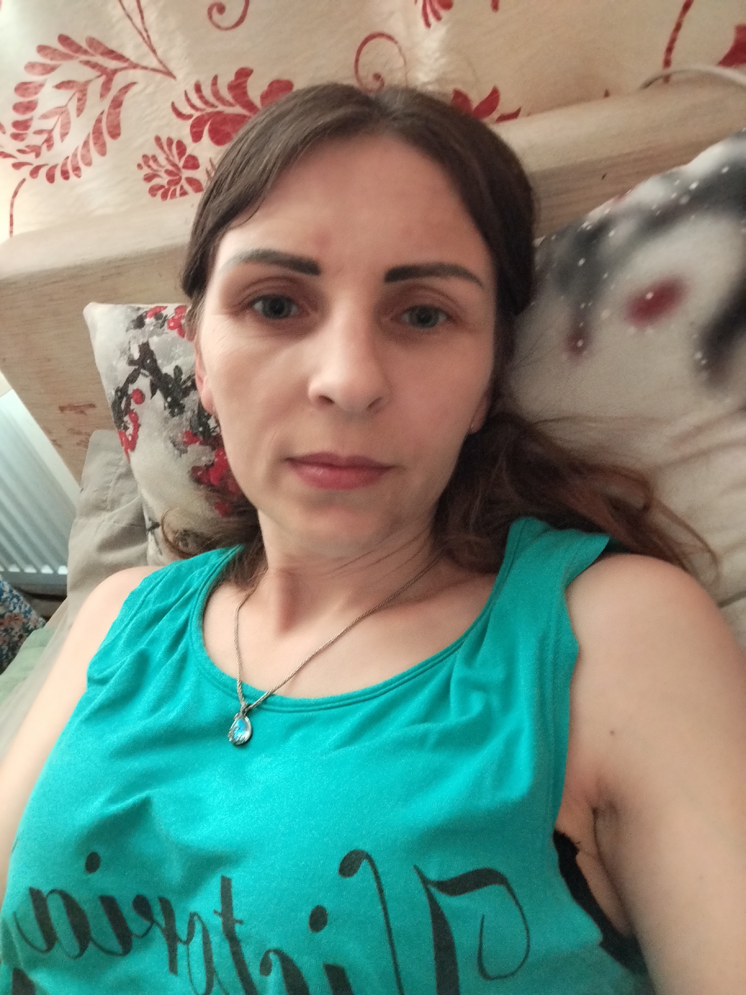 The image is a close-up selfie of a woman lying down, likely in a bedroom. The woman has brown hair and is wearing a turquoise tank top with a silver necklace that has a blue pendant. The background includes a patterned pillow and a headboard. The overall lighting is dim, suggesting it may have been taken indoors. There is no visible indication of the location being in Vinnytsia, Ukraine.