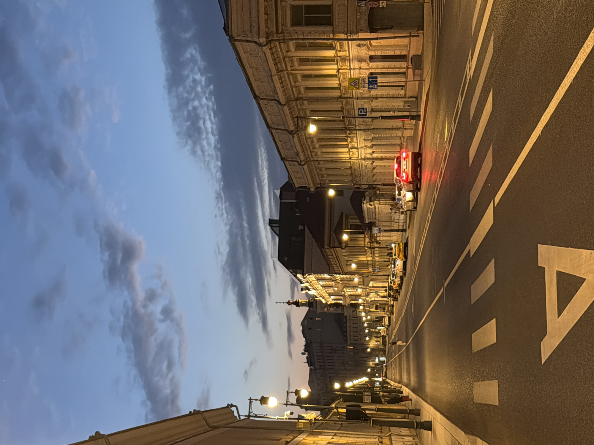 The image depicts a street scene in Moscow, Russia, at dusk. The sky is a deep blue with scattered clouds, and streetlights illuminate the asphalt road, which has clear white markings, including a crosswalk. Buildings line both sides of the street, some with classical architectural details and lit windows. Several cars are parked and driving on the road, including yellow taxis. A few pedestrians are visible on the sidewalks and crossing the street. The overall atmosphere is calm and urban.