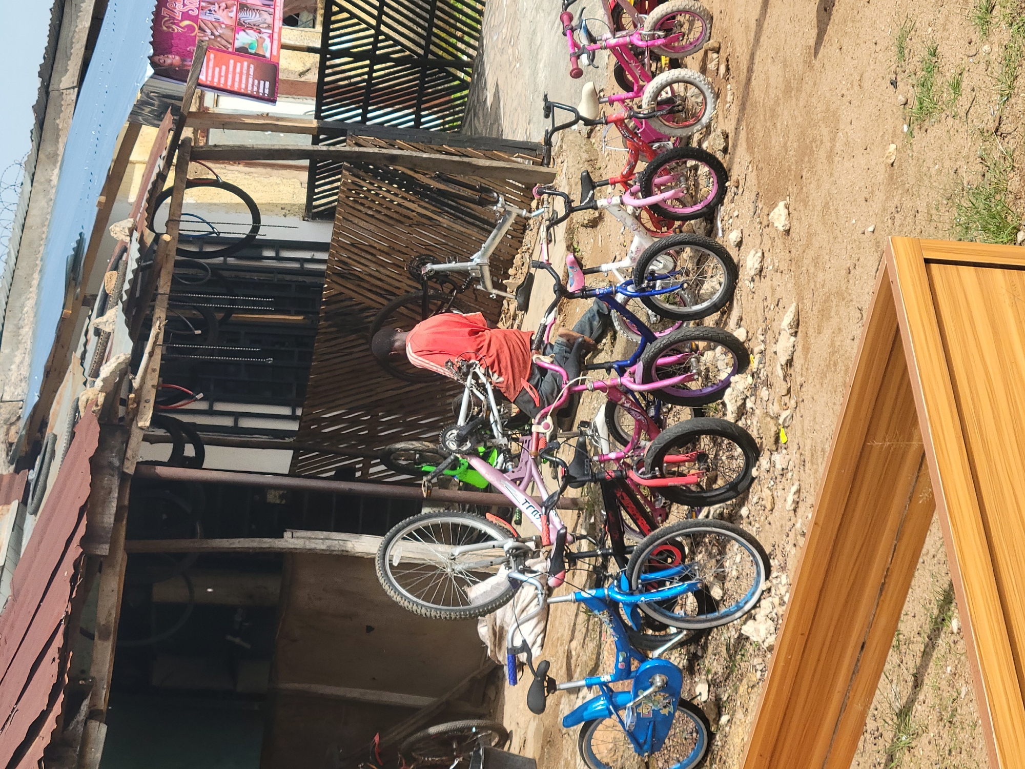 The image depicts an outdoor bicycle repair shop in Jos, Nigeria. A young person, wearing an orange t-shirt and jeans, is seated and appears to be working on a bicycle frame. A collection of bicycles, mostly children's bikes in various colors including pink, blue, and white, are arranged in front of the shop. The shop itself has a corrugated metal roof and wooden structures. In the background, a fence and a sign with images of hairstyles and nail services are visible. The ground is dirt with scattered rocks.