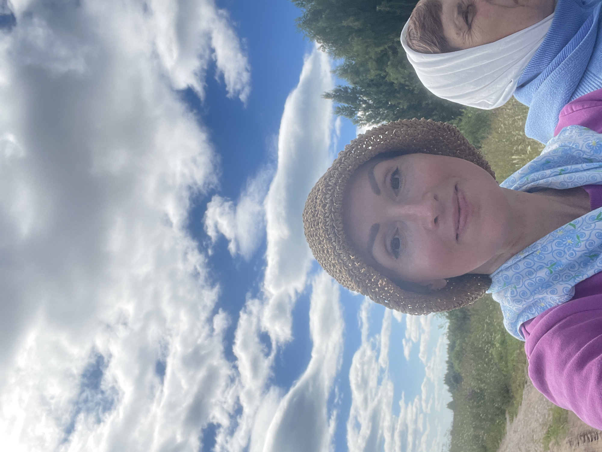 Two women are taking a selfie outdoors. The woman in the foreground on the left is wearing a straw hat, a pink sweatshirt, and a light blue scarf with a floral pattern. She is looking at the camera with a slight smile. To her right, slightly behind her, is another woman wearing a white headscarf and a blue top. She is looking off to the side. They are standing in a field with tall grass and some trees in the background. The sky is blue with white, fluffy clouds. This scene is located in the village of Дресвяново, Russia.