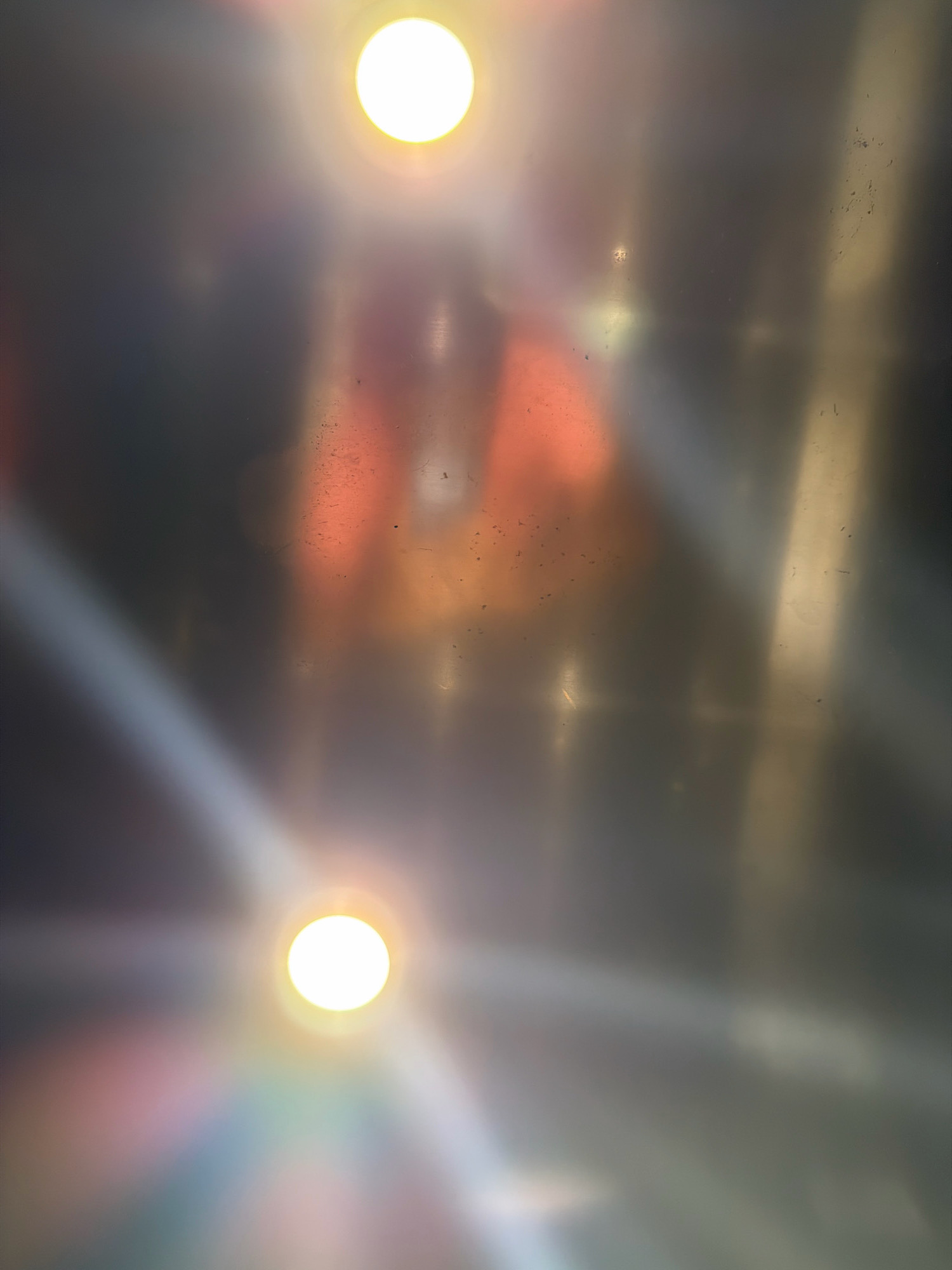 The image is a close-up, abstract shot of a metallic surface with lights reflecting off it. Two bright, circular lights are visible, one towards the top center and another towards the bottom center. Beams of light emanate from these sources, creating a diffused, ethereal effect. Streaks of color, possibly from further reflections or diffraction, appear near the bottom light. The metallic surface appears to be somewhat textured and shows minor imperfections, such as specks. The overall impression is one of dynamic light play and a somewhat industrial or utilitarian setting. This image does not contain any recognizable landmarks or features that would allow for specific identification of its location within Ayia Napa, Cyprus.
