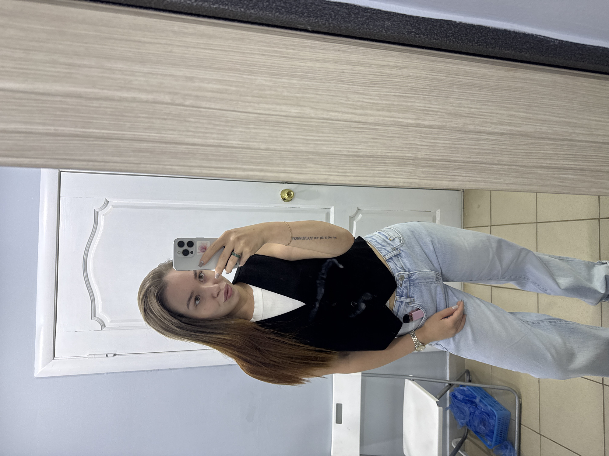 A young woman with long brown hair is taking a mirror selfie in what appears to be a dressing room or restroom setting. She is wearing light-wash jeans, a white t-shirt, and a black vest. She is holding a smartphone in her left hand, the camera pointed towards the mirror. On her left wrist, she wears a wristwatch, and there appears to be a tattoo on her left forearm.
The mirror reflects a portion of a room with light gray walls and a white door with decorative molding. The upper portion of the mirror frame is wood-grain patterned. The floor is tiled in a light tan color. In the foreground is a white chair and a blue plastic basket containing several disposable shoe covers.
Based on reverse image searches, the image is likely taken in Chaikovsky, Russia. The specific location within the city cannot be determined from the image.