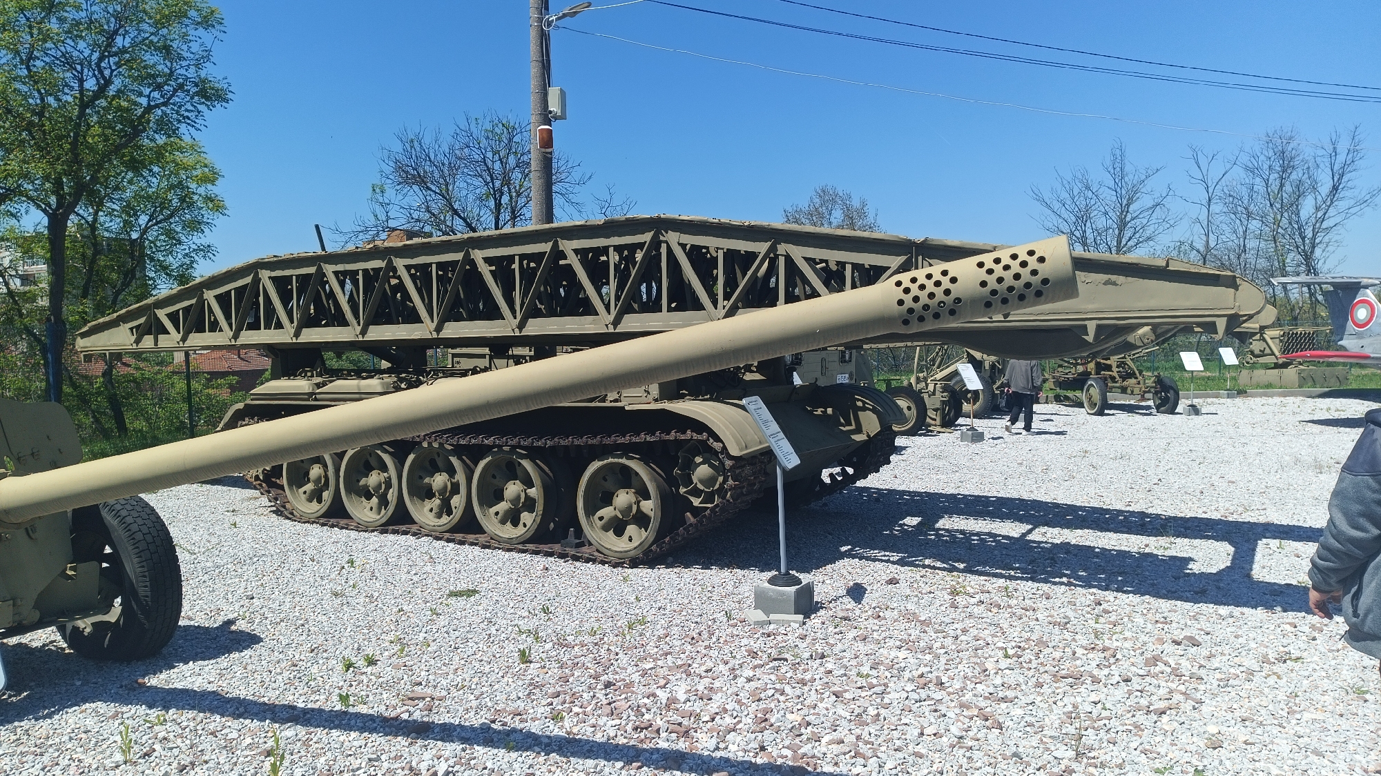 The image shows a military vehicle exhibit in Yambol, Bulgaria. A tank with a bridge-laying mechanism is the primary subject. An artillery cannon sits to the left, partially obscuring the tank. Several individuals are viewing the exhibit in the background. The exhibits are placed on a gravel ground. The weather is clear and sunny, as indicated by the blue sky. Bare trees and a building can be seen in the background.