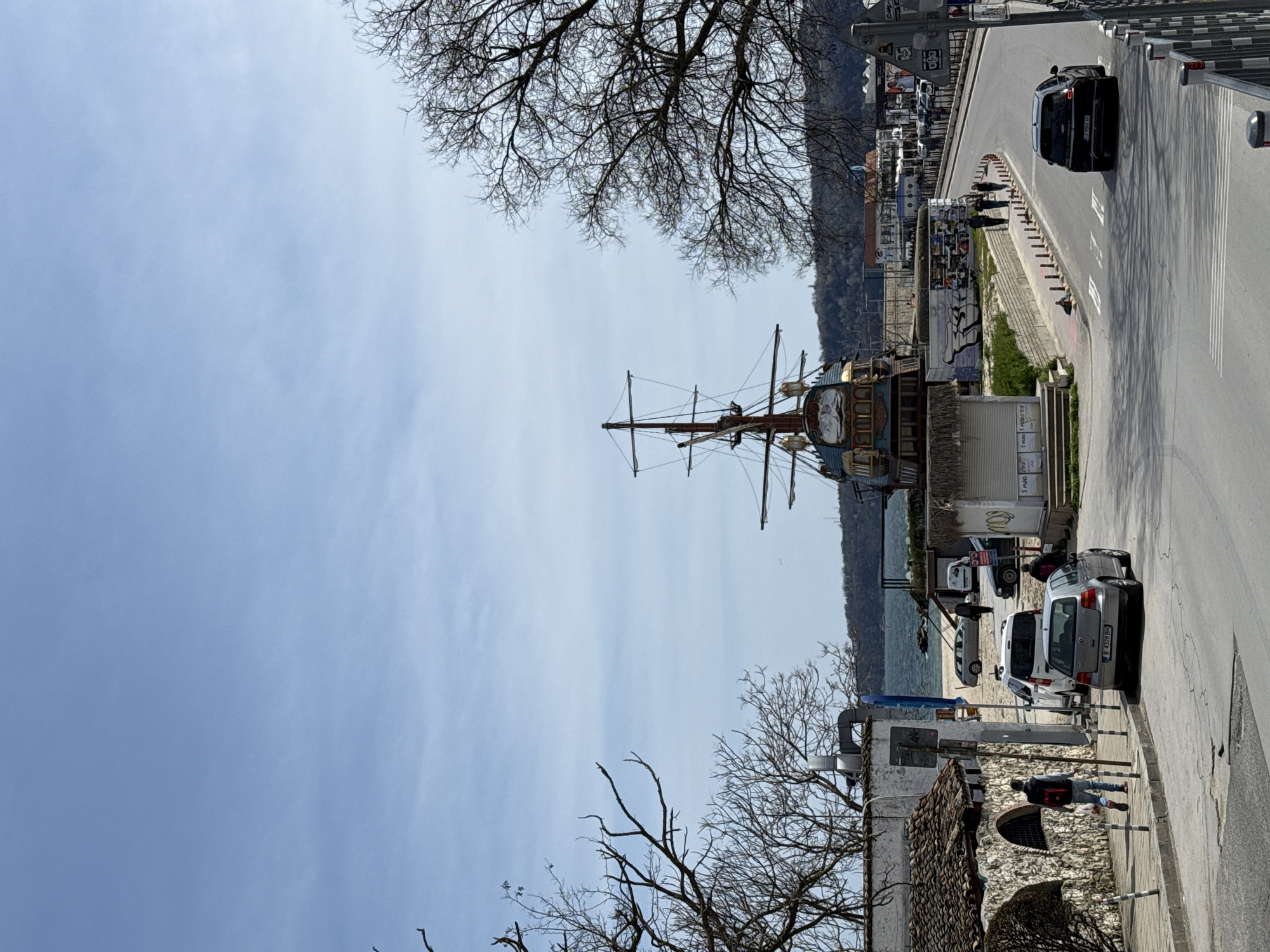 The image captures a street view in Varna, Bulgaria, featuring a prominent ship-shaped building with masts and nautical decor. The sky is partly cloudy. Bare trees frame the upper part of the scene. Along the right side of the image is a road with vehicles, including a dark-colored car moving along the road and parked cars closer to the building. Pedestrians are walking near the ship-shaped building and along the road. A stone structure with a tiled roof is situated at the lower right. In the background, a beach and the Black Sea are visible.