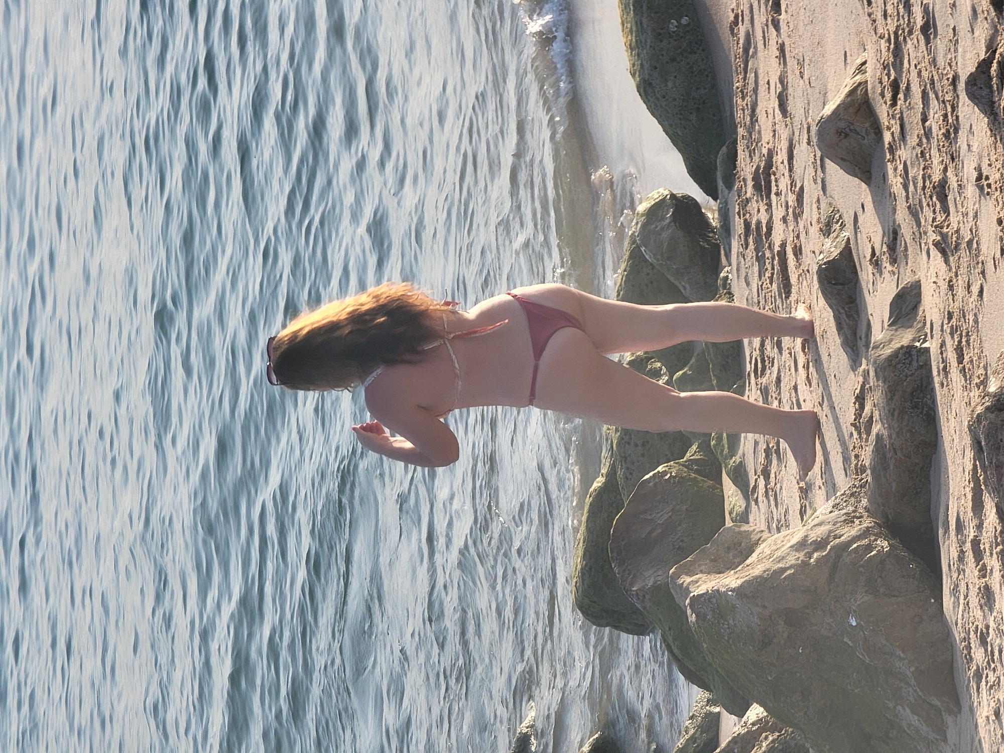 A woman wearing a burgundy bikini stands on a sandy beach with rocks in Quarteira, Portugal. She is facing away from the camera, with her hands raised to her face. The ocean is behind her, with waves gently lapping at the shore. The sand is wet in some areas, indicating the tide is coming in. The rocks are covered in green algae, suggesting they are often submerged in water. The sun is shining, casting long shadows across the beach.