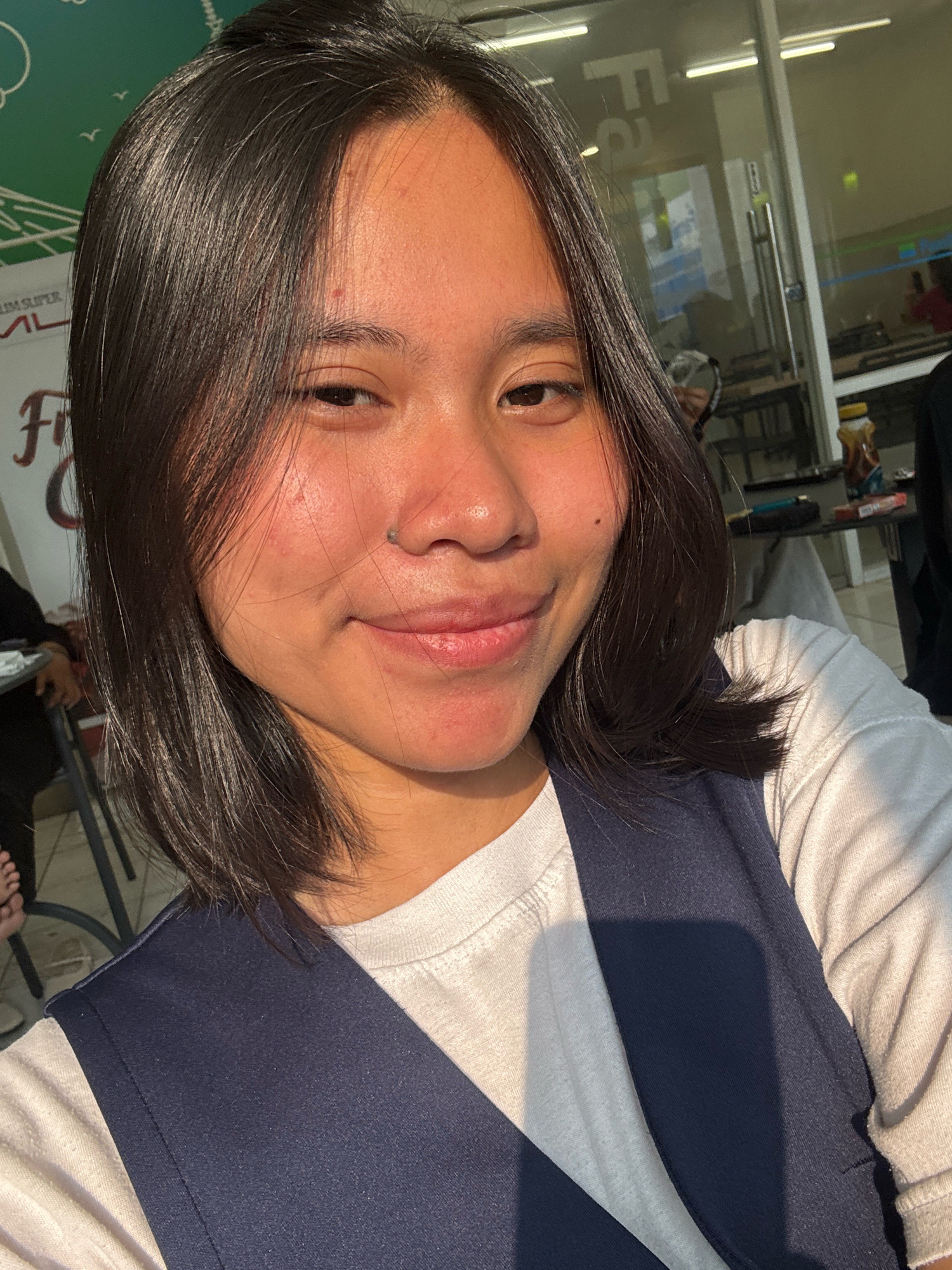 The image is a close-up selfie of a young East Asian woman with dark hair and a slight smile. She is wearing a white t-shirt and a dark blue vest. The lighting suggests it is either early morning or late afternoon, with sunlight casting a warm glow on her face. In the background, a glass-walled building with fluorescent lights is visible, along with tables and chairs, indicating an indoor or semi-outdoor setting that could be a cafe, restaurant, or office space. Part of a sign with "F2" is visible on the glass. The presence of a bottle of what appears to be honey or syrup on a table suggests a food or beverage establishment. Based on the context provided, this scene is located in East Jakarta, Indonesia.