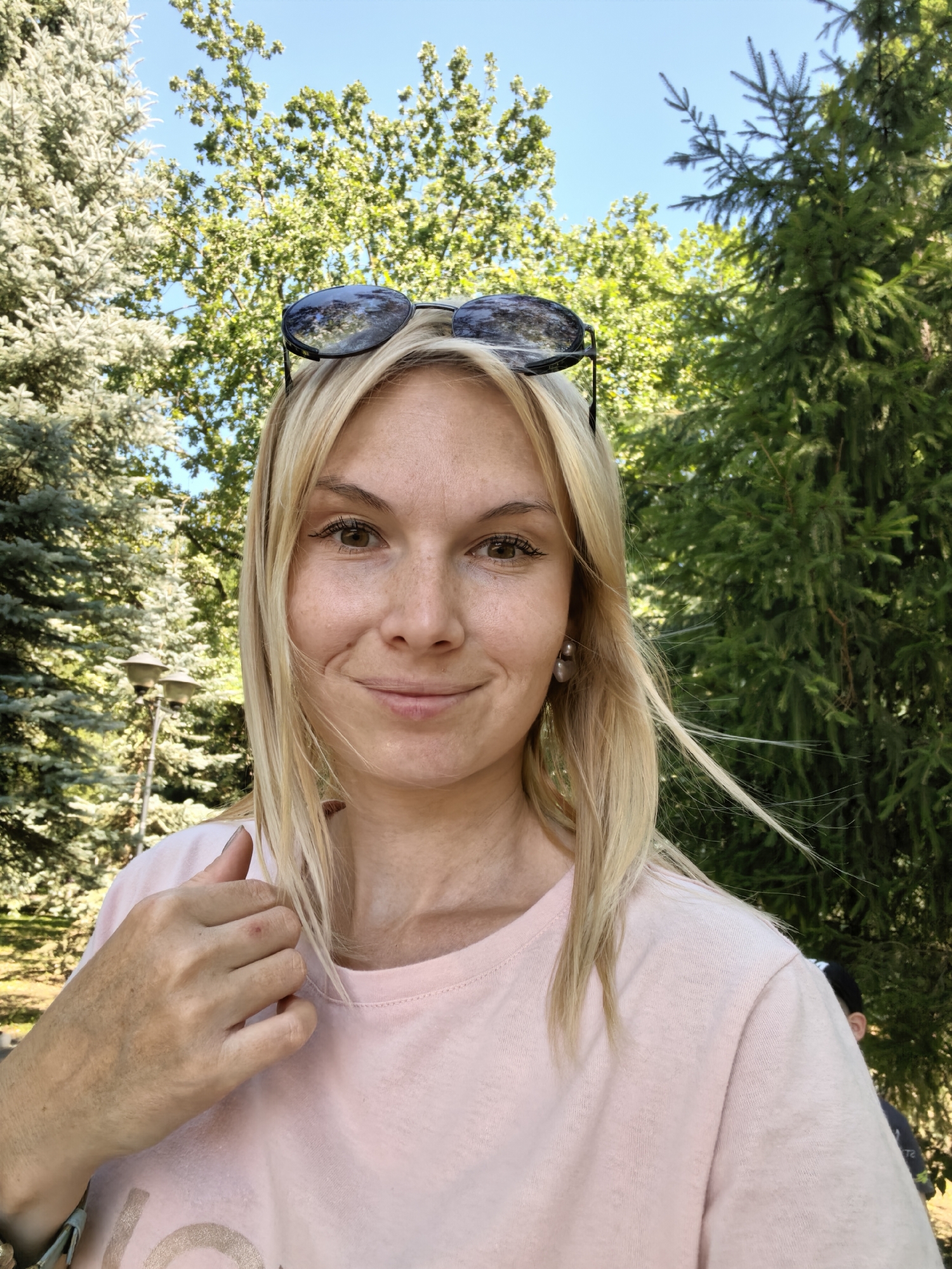 A blonde woman is taking a selfie outdoors. She is wearing a light pink t-shirt and sunglasses are perched on top of her head. Her hair is light blonde and falls around her shoulders. The background is filled with lush green trees and a clear blue sky. To the left of the woman, a lamppost is visible. The scene appears to be a park or a garden.