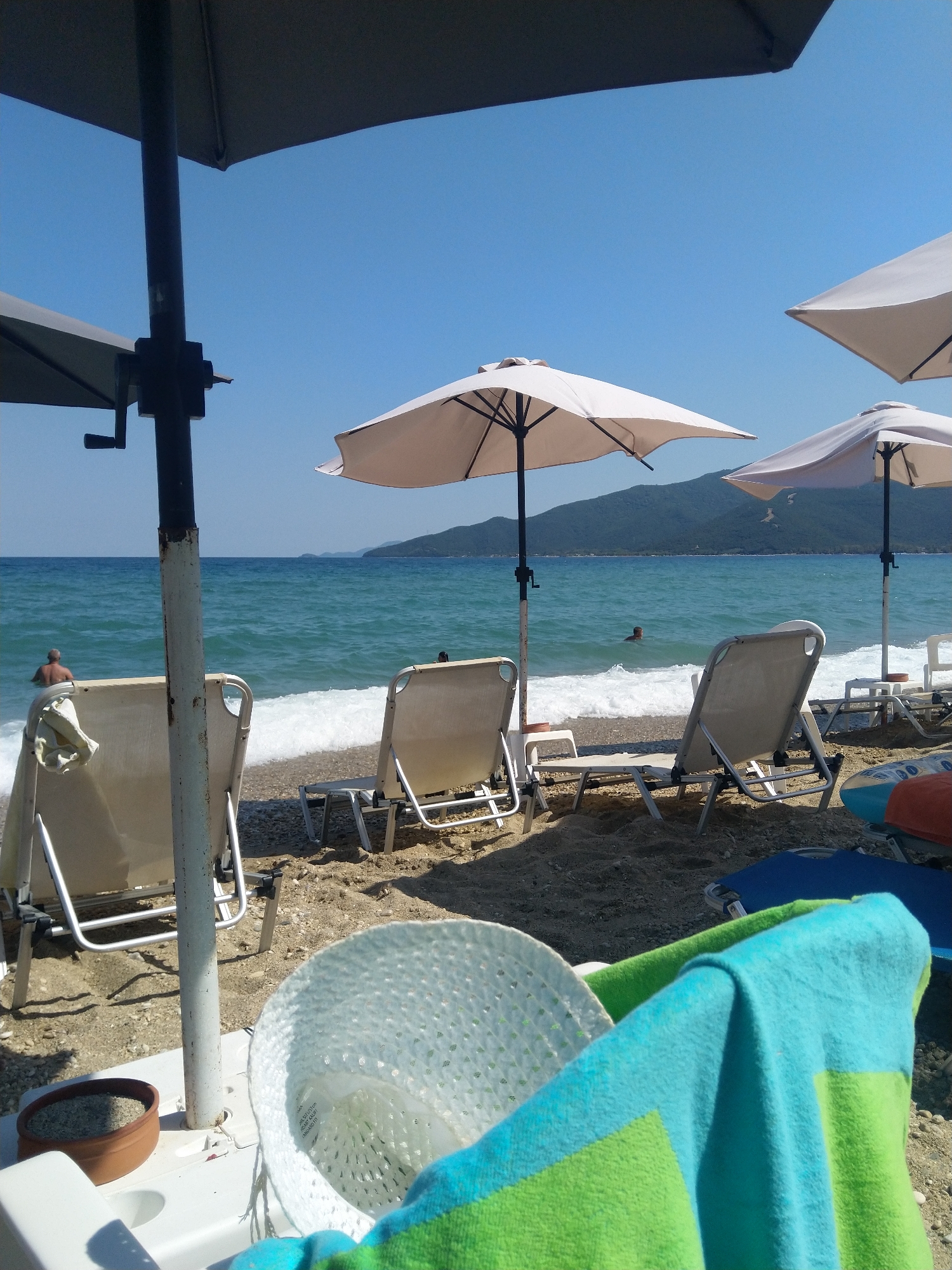 The image depicts a sunny day at a beach, likely in Nea Vrasna, Greece, given the presence of typical beach amenities and the mountainous coastline in the background. In the foreground, there are several empty lounge chairs set up on the sandy and pebbly shore, some partially obscured by beach umbrellas. A light-colored, woven hat is placed on a white plastic structure, next to a small terracotta pot containing what appears to be soil or sand. A vibrantly colored beach towel, with stripes of blue and green, is draped over the edge of a lounge chair in the right foreground.
The sea, with gentle waves breaking on the shore, stretches out towards the horizon. In the middle ground, a man with graying hair is partially submerged in the water, facing away from the camera. The clear blue sky above indicates fair weather. The overall scene conveys a relaxed atmosphere of a beach vacation.
