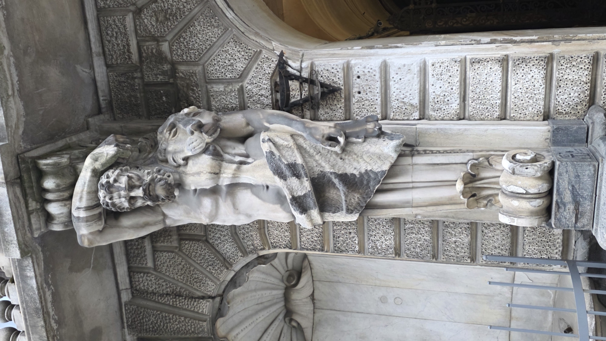 A close-up, medium shot of a stone architectural element, specifically a statue group, is displayed in Saint Petersburg, Russia. The statue appears to be of a man with a lion lying across him. The sculpture is detailed with carved elements around it. The surrounding architecture shows intricate designs and textures, including carved scrollwork and a textured patterned surface. A small piece of a railing is visible in the bottom right corner.