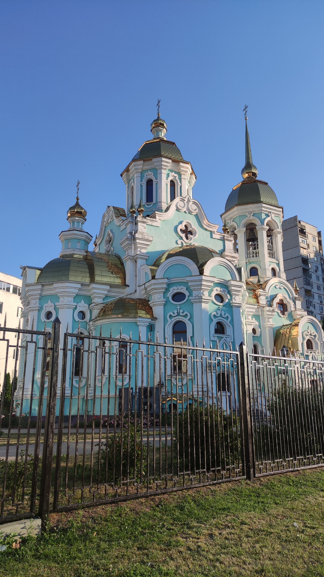 The image shows a view of an ornate church with a bright blue sky. The church features multiple golden domes and crosses, as well as light blue and white architectural elements. A tall, modern apartment building is visible in the background to the right of the church. In the foreground, a black metal fence with sharp finials runs horizontally across the image, partially obscuring the lower portion of the church and the green grass and bushes in front of it. The fence appears to be a barrier around the church property. The scene suggests a sunny day, likely in the late afternoon or early morning, given the angle of the light casting shadows. There are no people or specific activities depicted. The location is not explicitly identified, but the architectural style and the presence of the apartment building might suggest an urban or suburban setting in a region with Eastern Orthodox Christian influence. There is no visible text in the image.