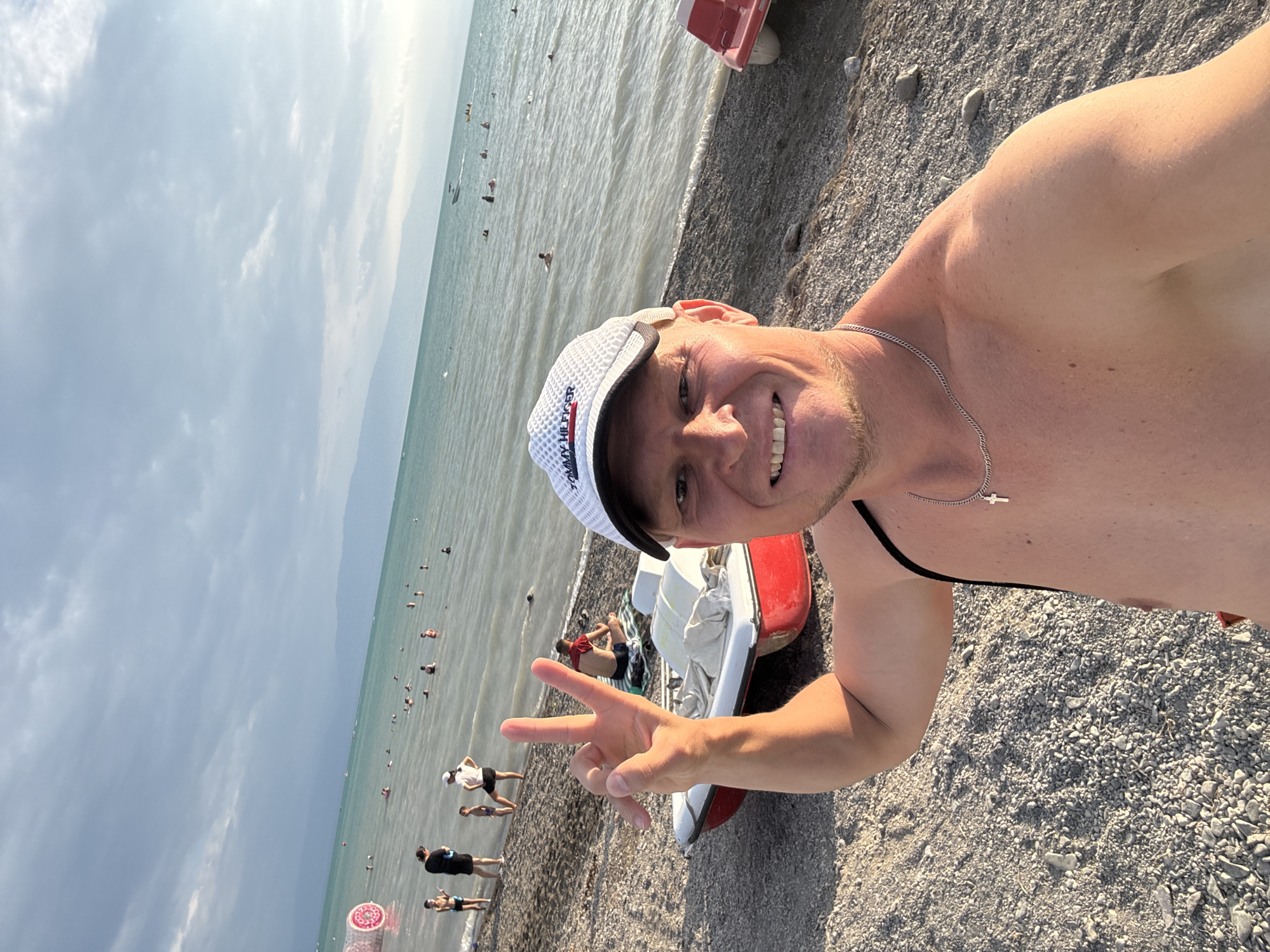 A man, wearing a white baseball cap with "Tommy Hilfiger" text and a silver chain with a cross pendant, smiles at the camera and holds up two fingers in a peace sign. He is on a pebble beach with the turquoise water of a lake behind him. In the water, many people are swimming and some are on paddle boats. There are mountains in the background. The scene is located in Zharbulak, Kazakhstan.