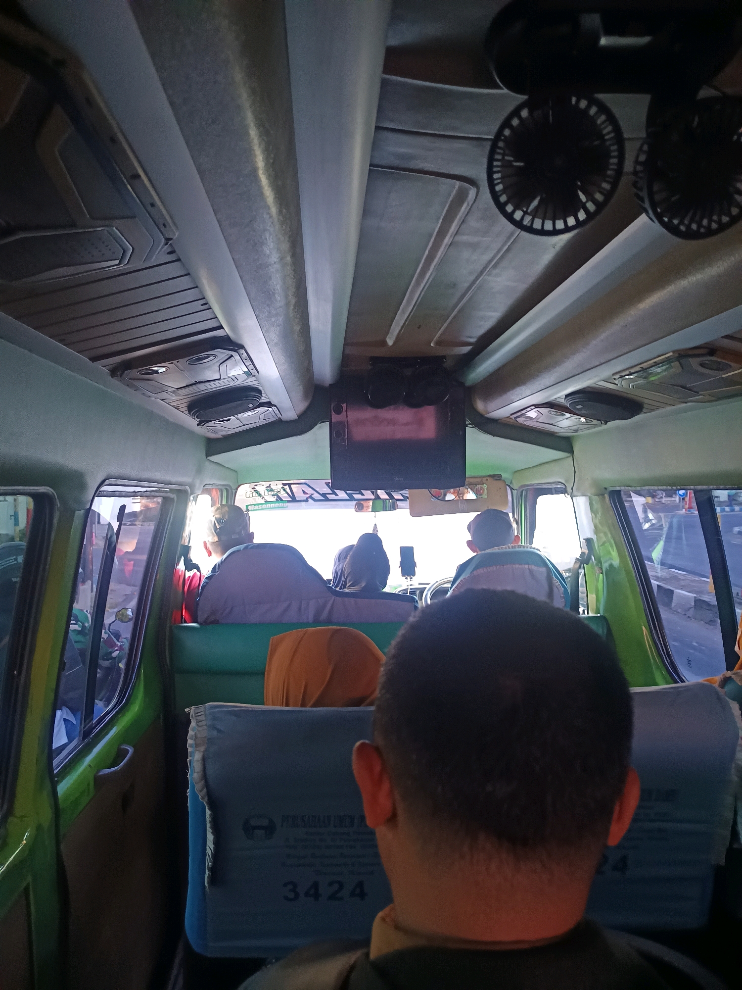 The image is taken from inside a green public transport vehicle, likely a minivans or small bus, looking towards the front and roof. Several passengers are visible, seated in the vehicle. The interior features light green upholstery, with some blue seat covers visible towards the rear. One seat cover has text and a number "3424" printed on it, suggesting it's a route or vehicle number. Above the passengers, the ceiling is detailed with ribbed panels and includes two small black fans and what appears to be a small screen or monitor. The windows of the vehicle offer a view of the exterior, showing traffic, including a motorcycle, and a street scene that is consistent with a location in Tanggumung, Indonesia. The overall impression is that of a typical journey within the city.