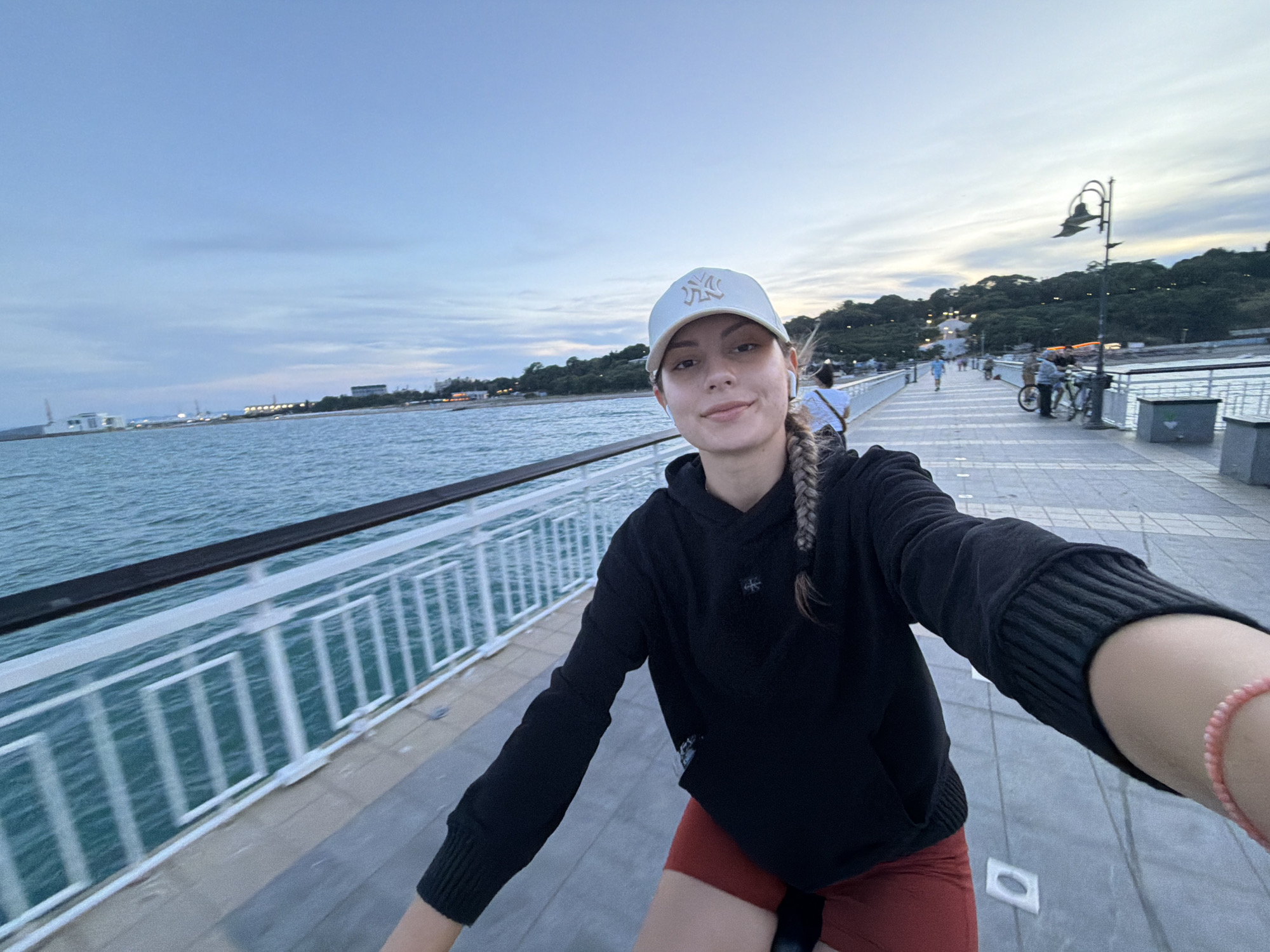 An eye-level, wide-angle selfie taken from a first-person perspective of a young woman with a braid and a white baseball cap, who is riding a bicycle along the Burgas Sea Garden Pier. The woman is wearing a black hoodie and red shorts. She is smiling at the camera. The pier, which is paved with light-colored square tiles, extends into the distance. A white railing lines the left side of the pier, separating it from the turquoise sea. In the background, there are trees and buildings on a hillside, and several people are walking and cycling on the pier. A lamppost with an ornate design stands on the right side of the pier. The sky is overcast with pale blue and grey clouds. The scene appears to be set in the late afternoon or early evening, based on the soft lighting.