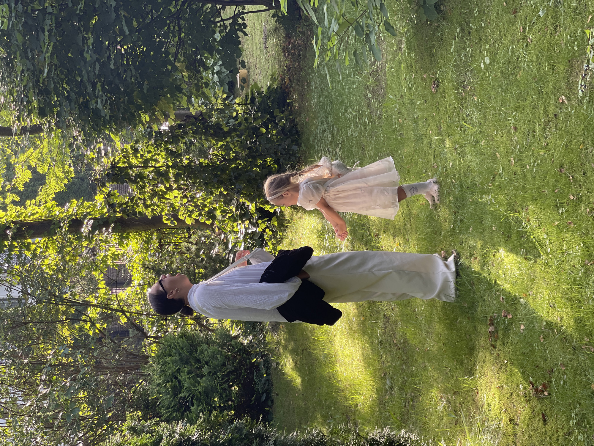 A woman and a young girl are standing on a grassy lawn in a garden or park. The woman is on the left, wearing a white shirt and light-colored pants, with a dark jacket draped over her arm. She is looking upwards and appears to be yawning or speaking. The girl is on the right, wearing a white dress and pink socks, and she is looking towards the woman with her hand outstretched. The environment is lush and green with trees and bushes surrounding the grassy area. Sunlight filters through the leaves, creating dappled light on the ground. The scene appears to be set in a tranquil outdoor location. The provided information indicates this location is in Svetlogorsk, Russia.
